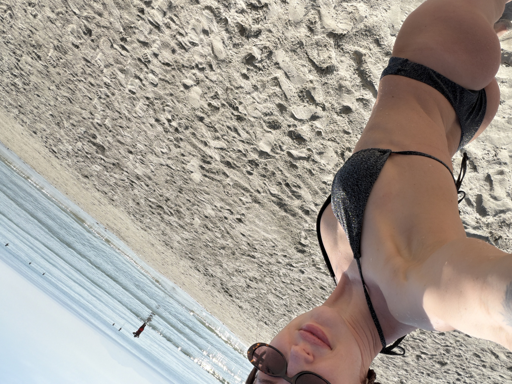 The image depicts a beach scene in Dong Ninh Hoa Ward, Vietnam, under clear daylight conditions. In the foreground, an adult individual, appearing to be female, is captured from a tilted, low-angle perspective, possibly a selfie. She is wearing a dark, glitter-textured two-piece bikini and sunglasses, with visible water droplets on her skin and a tattoo on her left forearm. The sandy beach dominates the mid-ground, showing numerous footprints. The ocean occupies the background, exhibiting calm, light blue waters with sunlight reflecting on the surface. Distant figures are visible in the water; one individual wearing a red garment is partially submerged, and several smaller, dark objects, possibly other people or buoys, are further out. The sky is light blue and clear.