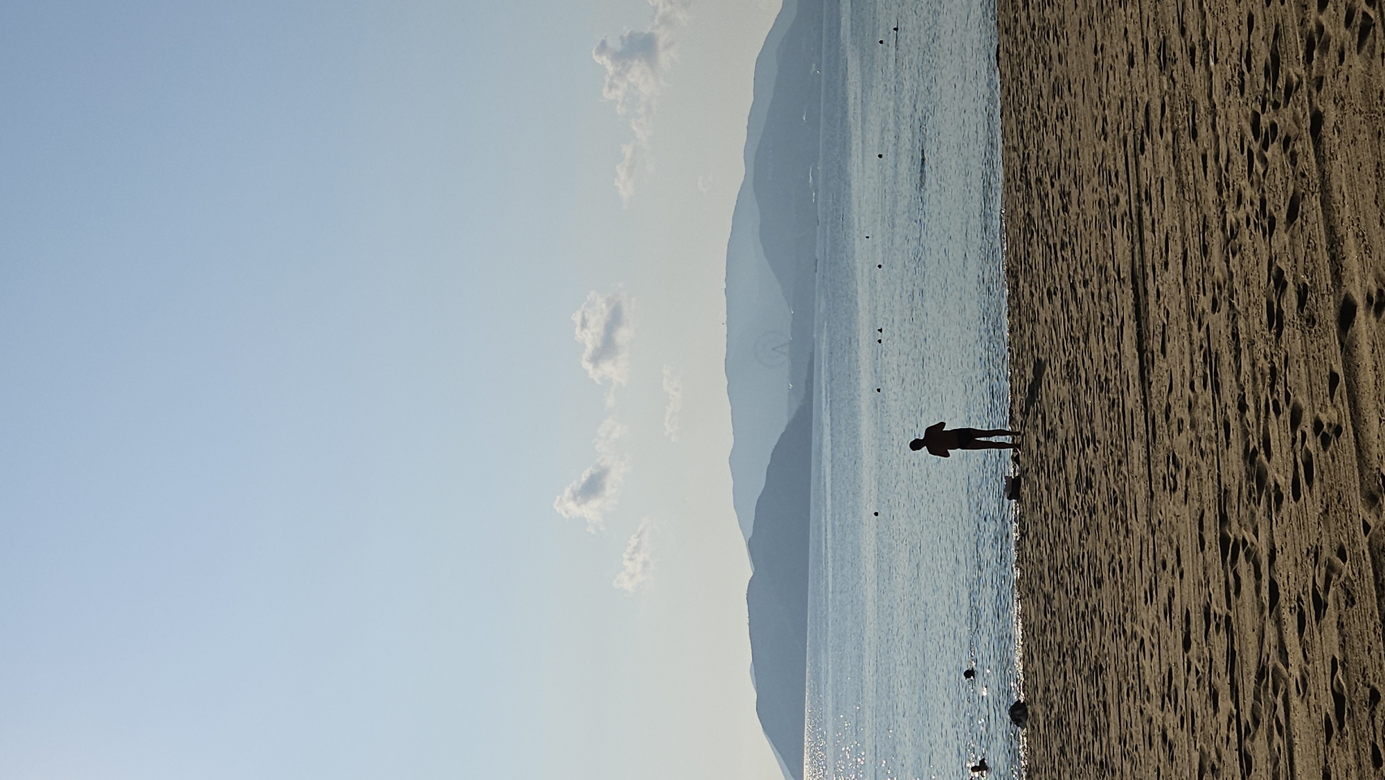 The image shows a beach scene, likely taken in Nha Trang, Vietnam, given the geographical context and the mention in the prompt. A person stands silhouetted on the sandy beach near the water's edge. The sand has numerous footprints. The sea extends towards the horizon, with faint outlines of mountains in the distance. The sky is clear and light blue, with scattered clouds. The overall scene is a coastal landscape, with the human figure as the central focus.
