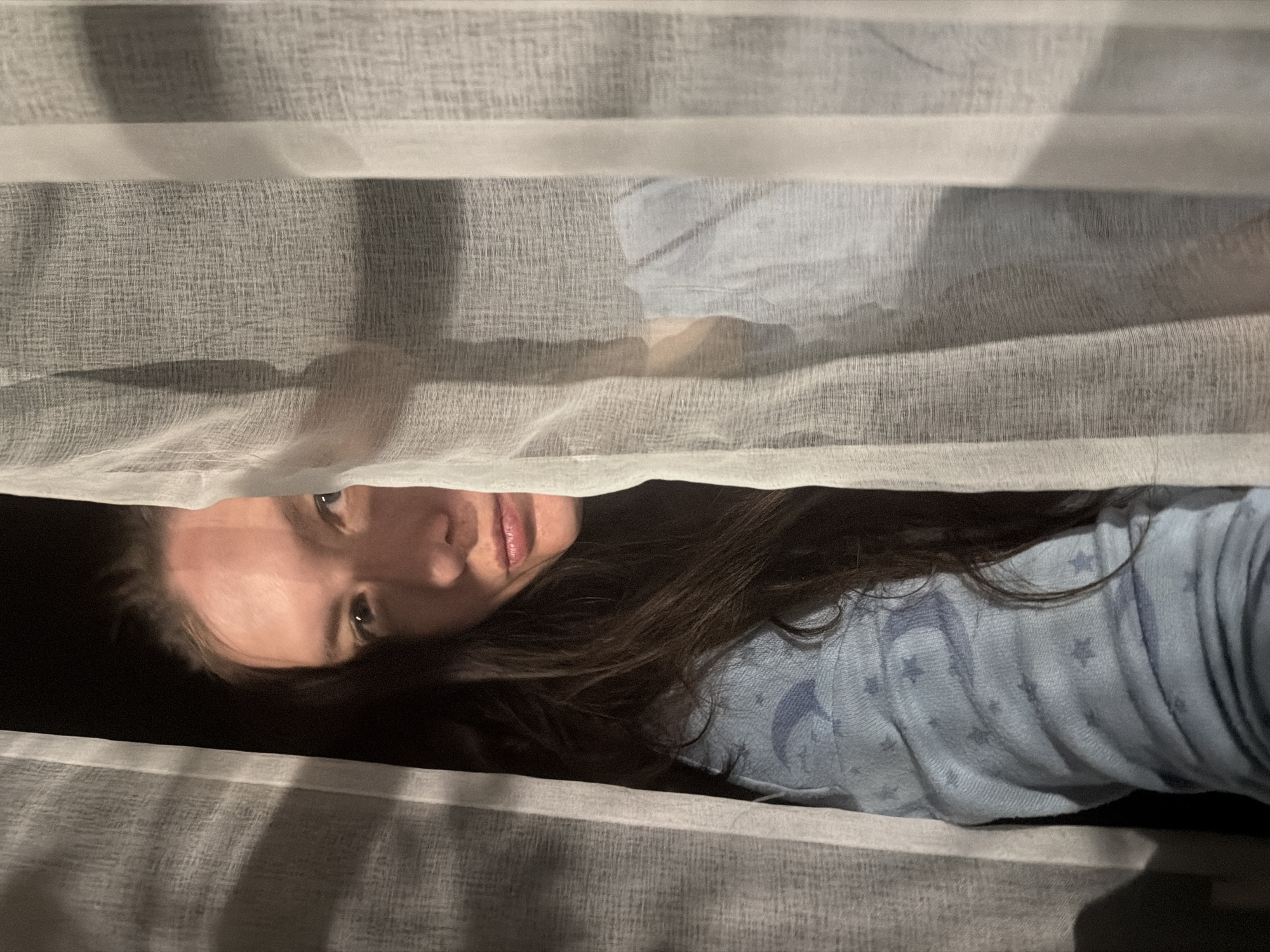 A woman is visible in an eye-level shot, partially obscured by light-colored, sheer curtains. The woman has dark hair and wears a light blue garment with star patterns. The background is dark, suggesting an interior space. The image is taken indoors with low lighting. The location of the photograph cannot be determined from the image provided, and there is no indication in the image that it was taken in the city of Staraya, Russia.