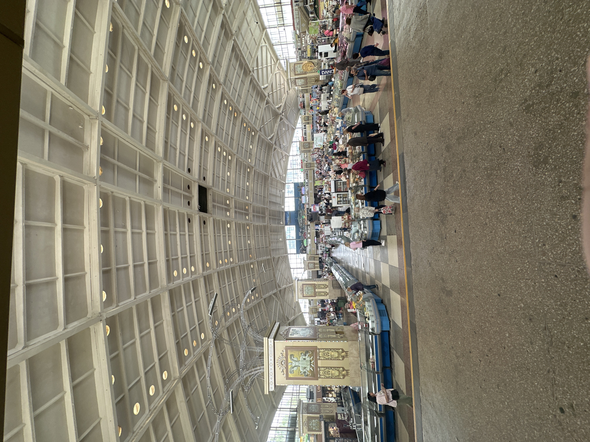 This image depicts the bustling interior of a large indoor market, likely a food market, located in Minsk, Belarus. The hall is characterized by an impressive, high arched ceiling constructed from a grid of light-colored beams, fitted with numerous circular recessed lights that cast a warm glow. Natural light also streams in from windows positioned along the upper sections of the arch.
Below this expansive ceiling, multiple rows of market stalls extend into the background. These stalls feature various display cases, many of which appear to be refrigerated, suggesting the sale of perishable goods. A diverse group of people, including both vendors and customers, are present throughout the market. They are seen standing, walking, browsing goods at the counters, and engaging in transactions, contributing to a lively atmosphere. Most individuals are casually dressed.
Prominent decorative elements include tall, ornate pillars adorned with intricate painted or tiled designs in shades of gold, brown, and blue. A large, complex metal sculpture or chandelier, featuring curving, cage-like structures, hangs from the ceiling in the central area of the market. The floor is a light-colored, speckled concrete or terrazzo surface, marked by a distinct yellow dividing line that delineates a walkway.
Visible text on a sign near one of the stalls appears to read "DANONE" or "DANO," hinting at a vendor selling dairy products. The scene takes place during daylight hours, likely late morning or afternoon, given the balance of natural and artificial illumination. The overall impression is one of an active and well-maintained public market.