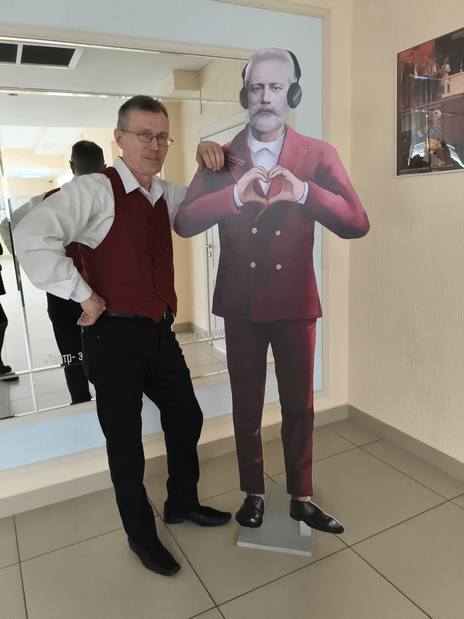 A man stands to the left of a cardboard cutout of Pyotr Ilyich Tchaikovsky. The man is wearing glasses, a white button-down shirt, a maroon vest, and black pants. He is posing with his left hand on his hip and his right arm around the cutout. The cutout is wearing a maroon suit, a white shirt, and headphones, and is holding his hands up to form a heart shape. The background is a wall with a mirror and a poster. The floor is tiled.