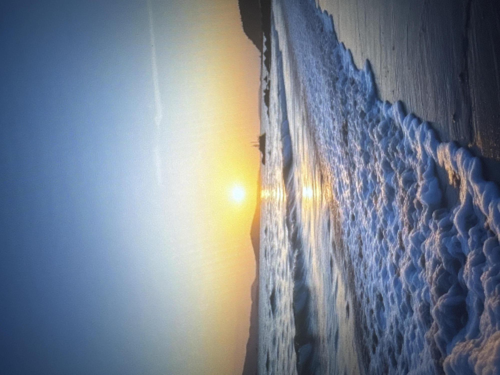 The image depicts a serene beach scene at sunset. The sun, a bright orb of golden light, is positioned low on the horizon, casting a warm glow across the sky and reflecting on the water. Gentle waves roll onto the shore, creating white foam that stretches across the sand. In the distance, silhouetted hills or mountains form a hazy backdrop. The overall atmosphere is peaceful and tranquil. This scene is not located in the city of Tyumen, Russia, as Tyumen is an inland city with no coastline.