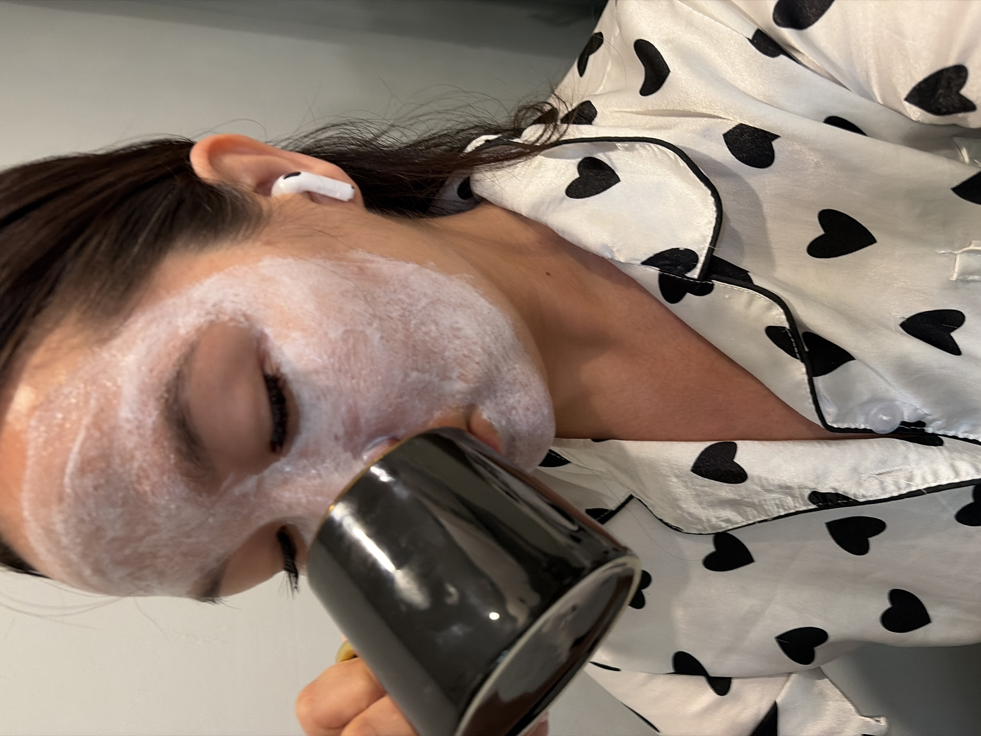 A woman with a face mask on her face is drinking from a black mug. She is wearing a white pajama top with black hearts on it and an AirPod in her right ear. The image appears to be taken indoors, with a neutral-colored wall in the background. There is no discernible indication of the city of Chaikovsky, Russia, or any specific location within it.