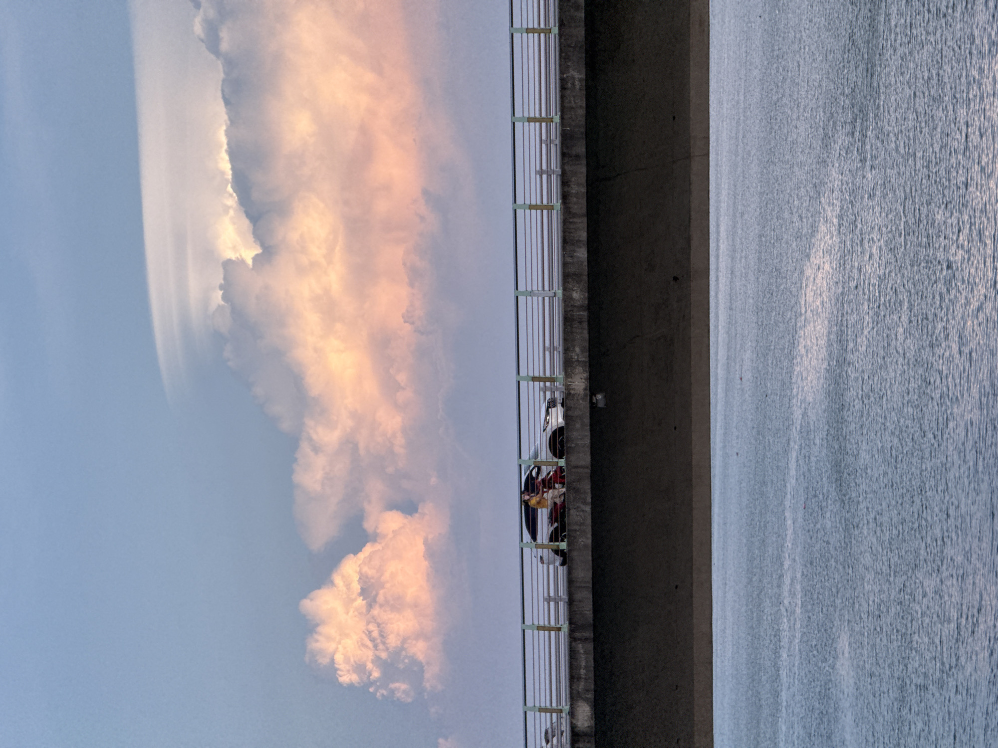 The image depicts a scene from a bridge in Da Nang, Vietnam, overlooking the Han River. The sky is filled with dramatic clouds tinged with pink and orange hues, suggesting either sunrise or sunset. A concrete bridge with metal railings stretches horizontally across the frame. On the bridge, a white SUV is visible, and next to it, a person is riding a red scooter. The scooter carries two occupants; the rider is wearing a yellow shirt, and a passenger is seated behind them. Below the bridge, the Han River appears calm, with ripples on its surface reflecting the ambient light.