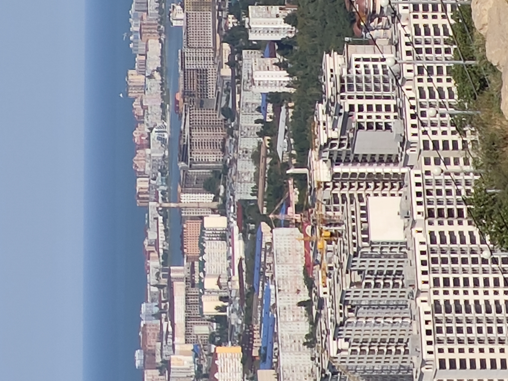 The image displays a panoramic view of a coastal city, likely Makhachkala, Russia, characterized by a dense urban landscape situated between the sea and rolling hills. In the foreground, a complex of modern, multi-story apartment buildings under construction dominates the scene. These buildings are characterized by their repetitive geometric facades, numerous windows, and visible structural elements indicating ongoing development, including cranes.
Further into the city, a mix of older and newer residential and commercial buildings are visible, varying in height and architectural style, painted in muted tones of beige, white, and pink. Some buildings feature balconies and distinct rooflines.
In the background, a body of water, presumably the Caspian Sea, stretches across the horizon, meeting a clear, pale blue sky. The overall impression is one of urban expansion and development in a seaside setting. The lighting suggests a bright, sunny day. No people are discernible in the image.