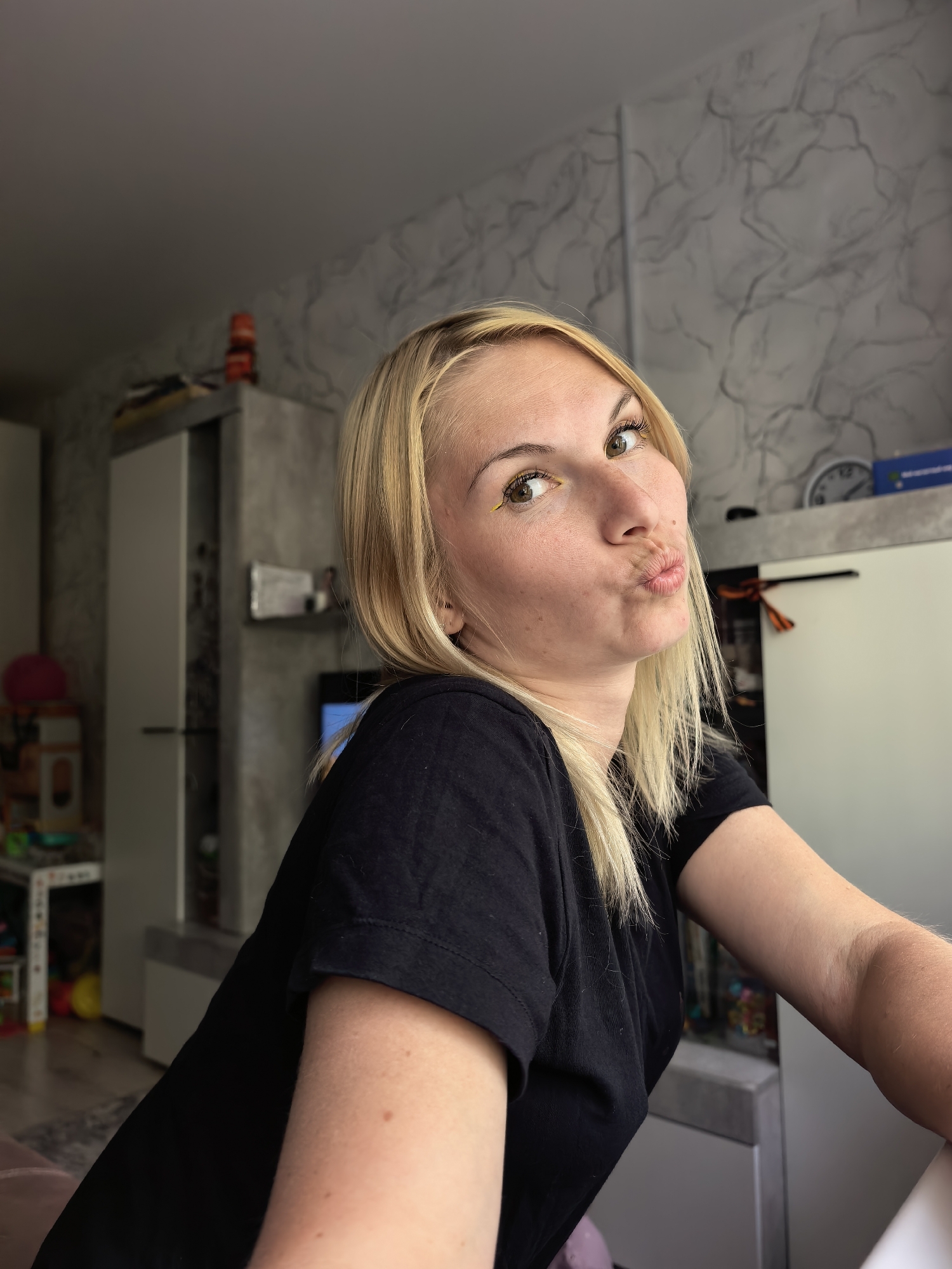 A young Caucasian woman with blonde hair and green eyes is captured in a close-up shot, puckering her lips for a kiss. She is wearing a black t-shirt. Her eyes are looking towards the camera, and she has applied yellow eyeliner. The background appears to be an indoor setting, likely a room in a residential building, with furniture and decor visible, including what looks like a wardrobe and a wall with a marbled texture. The lighting suggests it is daytime. The image was taken in Sterlitamak, Russia.