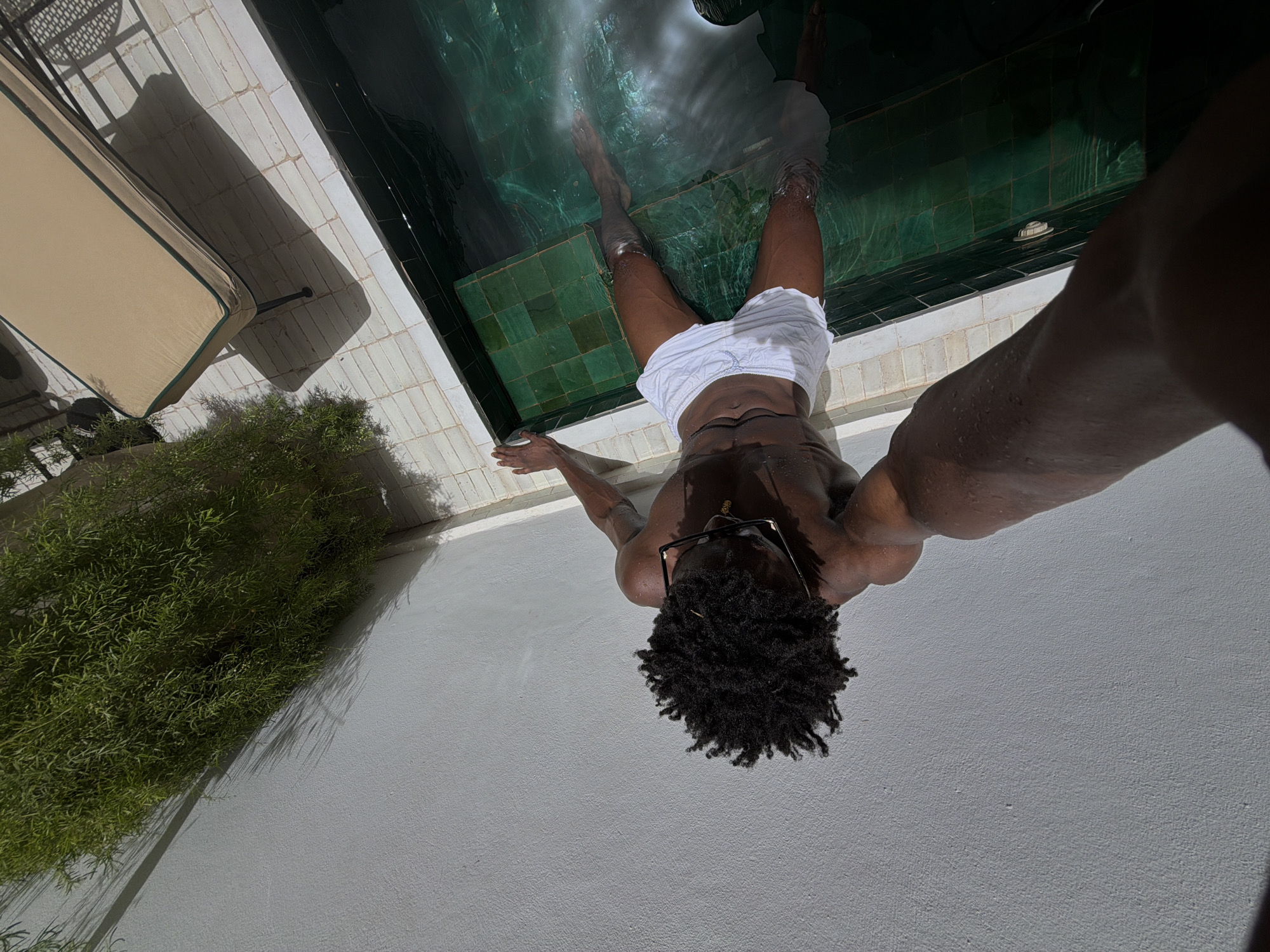 A man with dark skin and dreadlocks, wearing white swim trunks and glasses, is partially submerged in a rectangular pool with green tiled walls. He is lying on his back with his arms outstretched, his legs extended in the water. The pool is surrounded by a white tiled patio and a white wall. To the left of the pool, lush green foliage hangs over the wall, casting shadows. In the background, a light-colored lounge chair with a dark frame is visible. The scene appears to be an outdoor setting, possibly a courtyard or a private residence in Marrakech, Morocco, given the architectural style and lush vegetation often found in riads. The lighting suggests a sunny day.