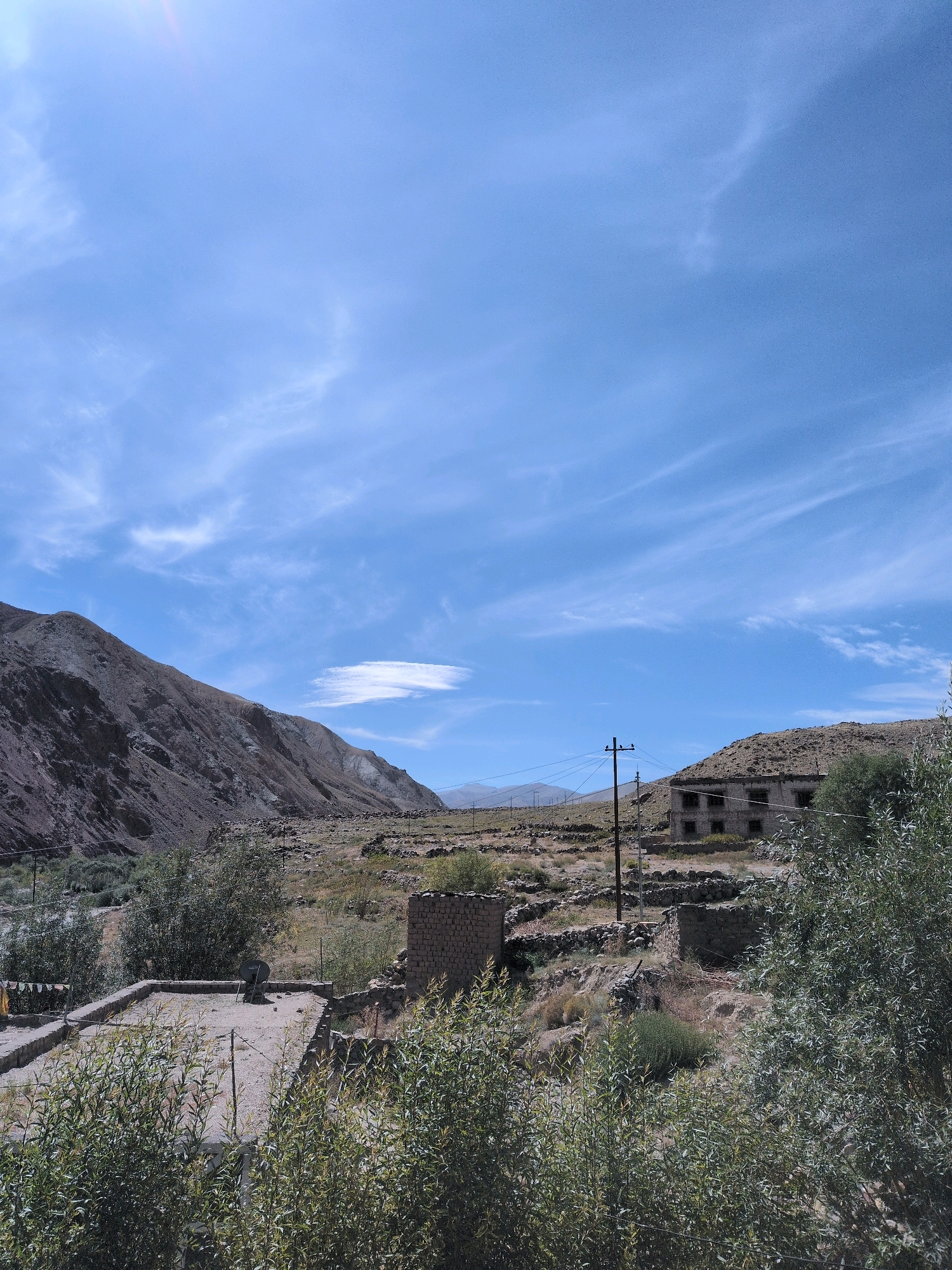 This image depicts a rural landscape with mountainous terrain and sparse vegetation, possibly in the vicinity of Lato, India. The sky is clear blue with wispy white clouds. In the foreground, there is a flat-topped structure, possibly a roof, with some debris and a satellite dish. Surrounding it are various types of greenery, including shrubs and trees.
Further back, there are more stone structures, some appearing to be low walls or foundations, and a few brick buildings. One prominent building is a multi-story structure with dark, empty window openings, suggesting it might be abandoned or unfinished. A wooden utility pole with wires stands near this building, and a few birds are perched on it. The land is dry and appears to be hilly or mountainous, with rocky slopes dominating the background. The overall scene conveys a sense of a remote, possibly underdeveloped area with a rugged natural environment.