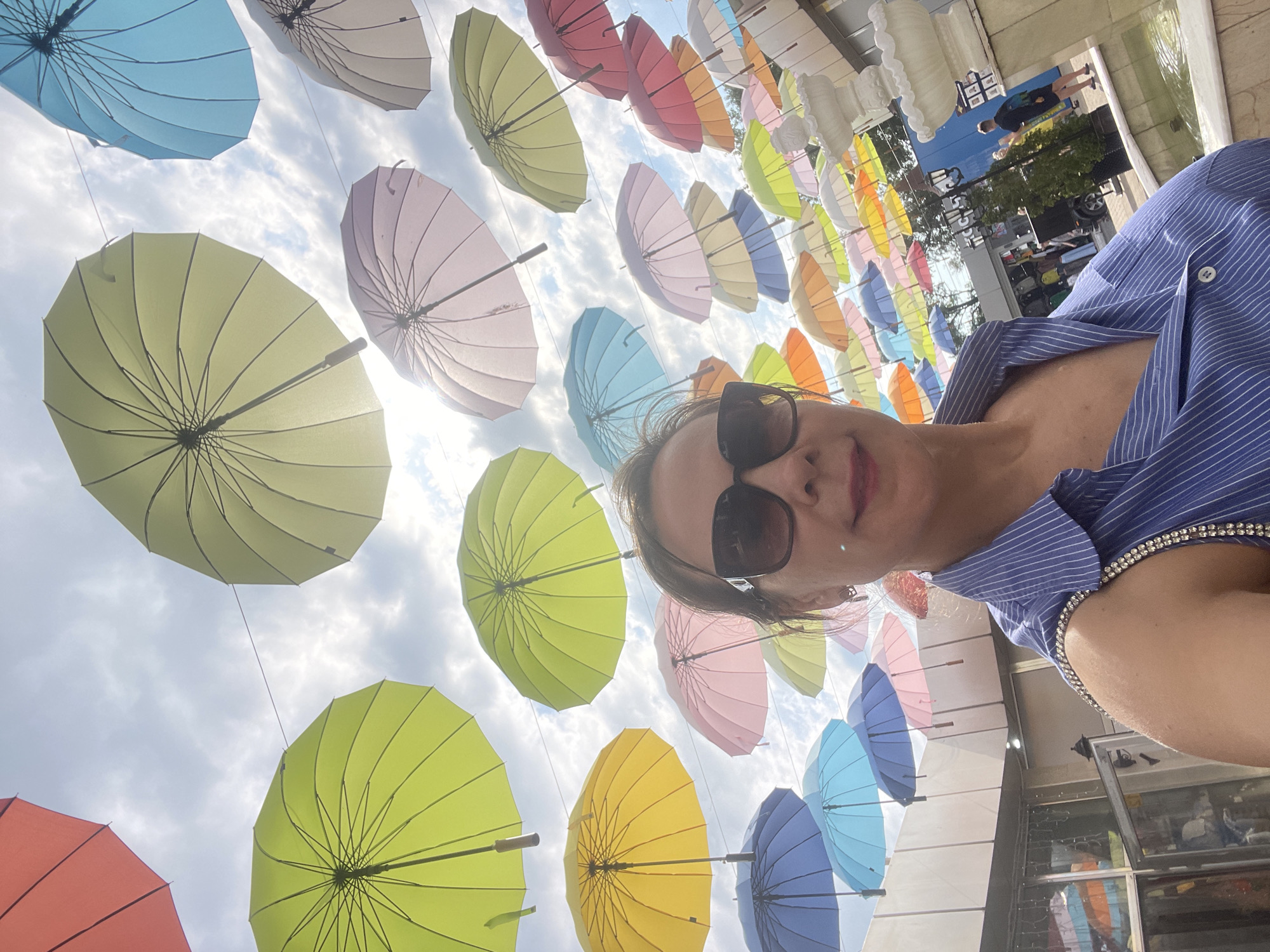 A woman takes a selfie outdoors, with many colorful umbrellas suspended in the sky above her. She is wearing a blue and white striped shirt and dark sunglasses. In the background, there are buildings, trees, and a fountain. This scene is located in Sochi, Russia.