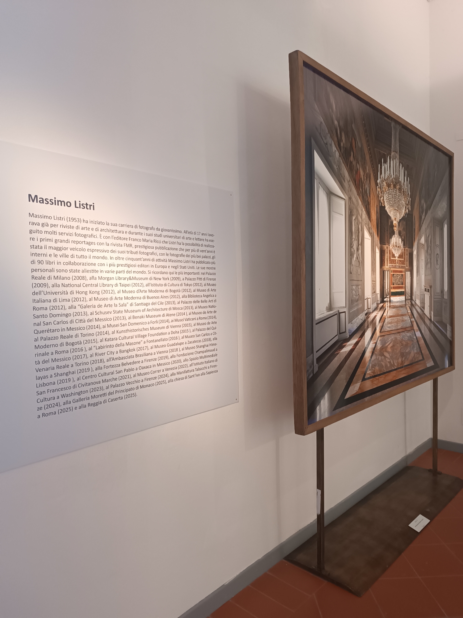 The image shows a photograph on display in an art gallery. The photograph depicts a long, ornate corridor with chandeliers, marble floors, and classical architectural details. The walls are decorated with paintings or reliefs. The perspective of the corridor suggests a grand palace or museum. The photograph is framed with a wooden border and mounted on a metal stand. To the left of the photograph is a wall with text describing the artist Massimo Listri and his career. The text mentions various exhibitions of Listri's work. The floor is made of brick. The image appears to be taken inside an art gallery, with white walls and a focus on the displayed artwork. There is no mention of the specific city of Viadana, Italy, in the provided information.