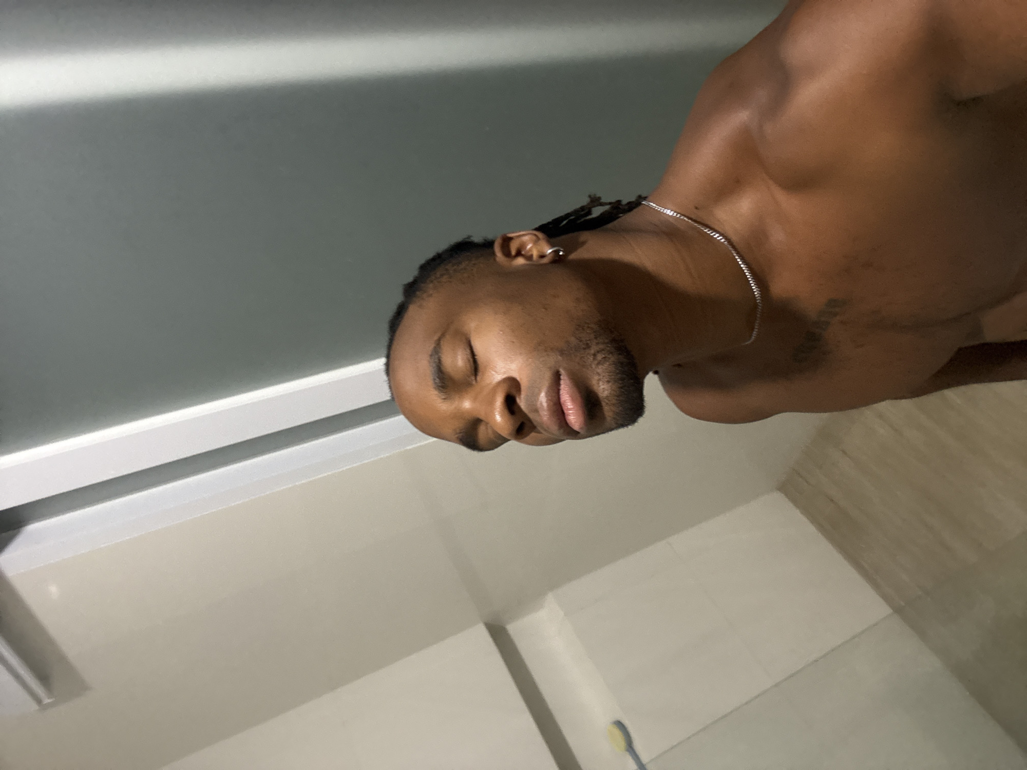 A medium-shot, low-angle, indoor photograph depicts a muscular, dark-skinned man with braided hair, an earring, and a thin silver chain necklace. His eyes are closed, and he has a short beard. A tattoo is visible on his chest. The background consists of bathroom tiles and a white window frame. The location is in a bathroom, in the city of Unknown, Unknown.
