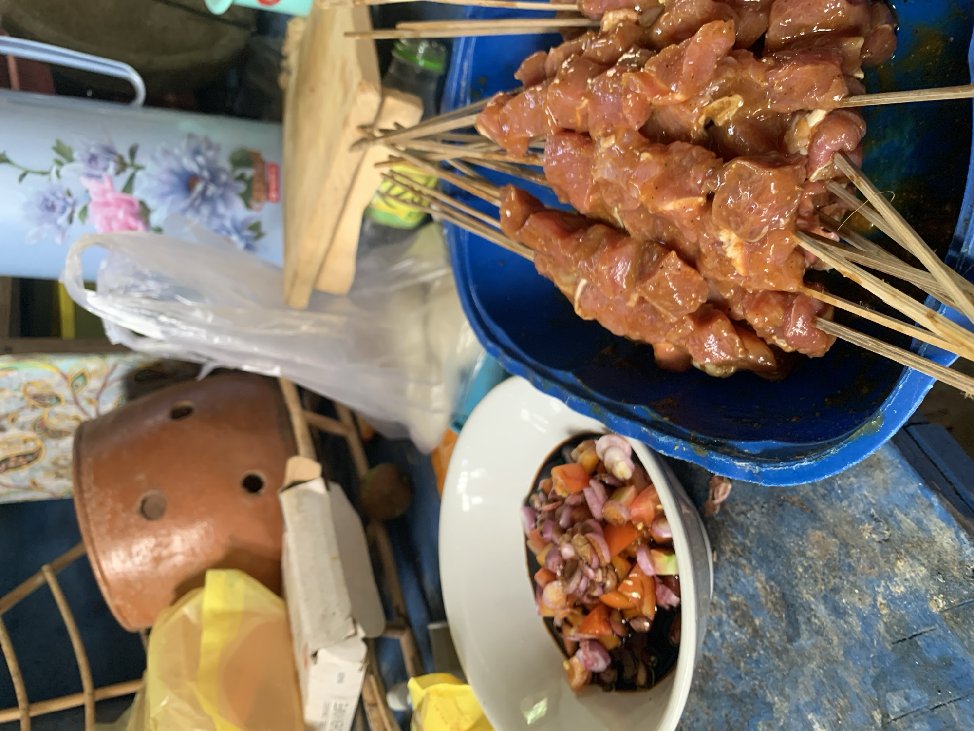 A close-up shot shows a collection of food items arranged on a possibly makeshift surface in Karangkemiri, Indonesia. Dominating the upper right is a blue bowl filled with skewers of seasoned, raw meat. Adjacent to this is a white bowl containing a mixture of chopped ingredients in a dark sauce, appearing to be a relish or dipping sauce.
In the background, a decorated container with a floral pattern sits alongside a wooden cutting board and a green bottle. A brown ceramic object with holes is visible amidst other items, including pieces of cardboard and plastic wrap. The objects rest on a blue surface with worn paint.