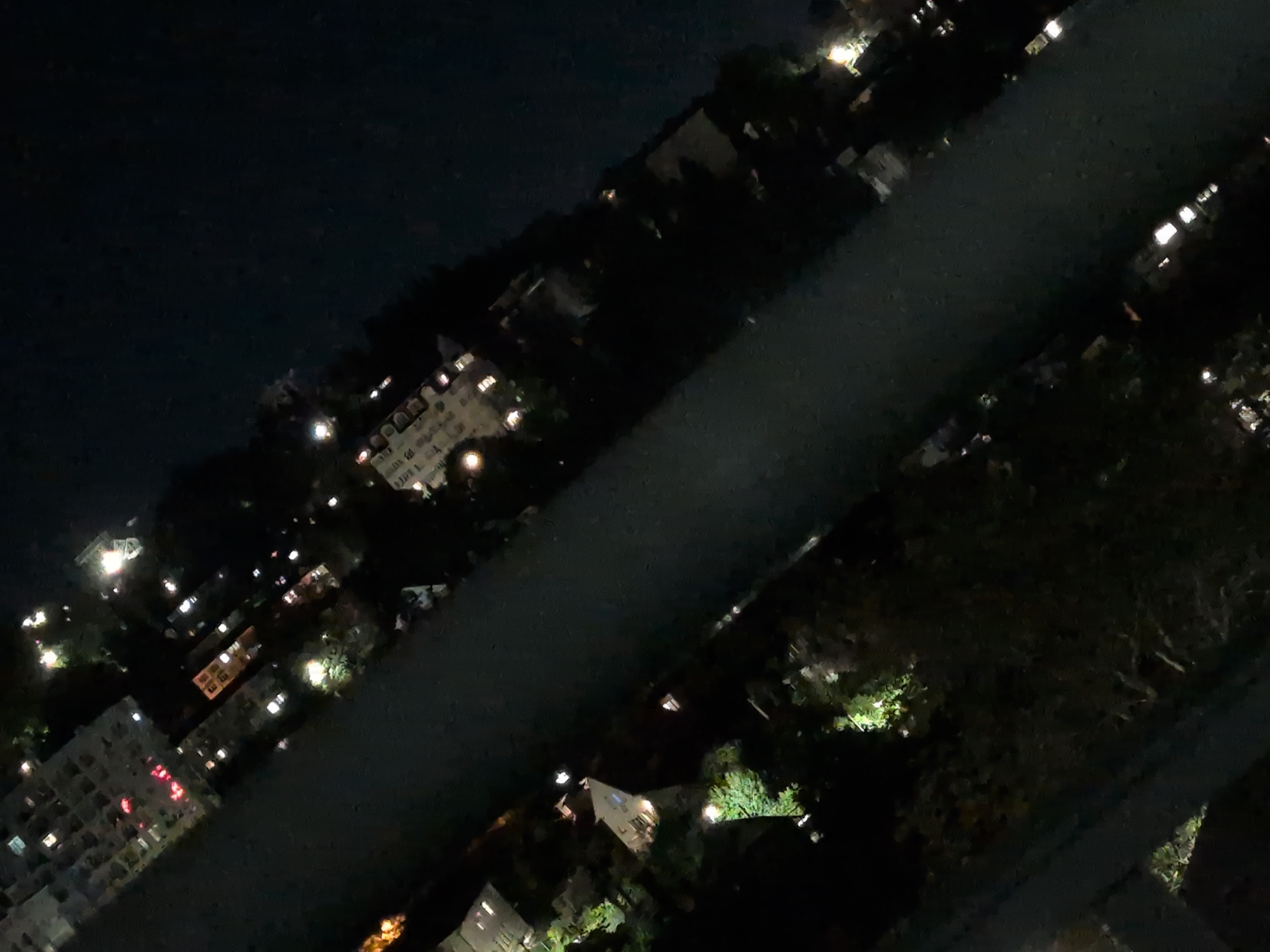 An aerial, night-time photograph captures a section of the city of Sochi, Russia, with a body of dark water, likely a river or canal, bisecting the scene. The water reflects some of the scattered lights from the surrounding urban and residential areas. On either side of the water, buildings and trees are visible, illuminated by artificial lights emanating from windows and street lamps. The perspective is tilted, giving the impression of looking down at an angle. The overall ambiance is one of a quiet, sleeping city. There are no people or specific actions clearly depicted, only the static presence of infrastructure and vegetation under the cover of darkness.