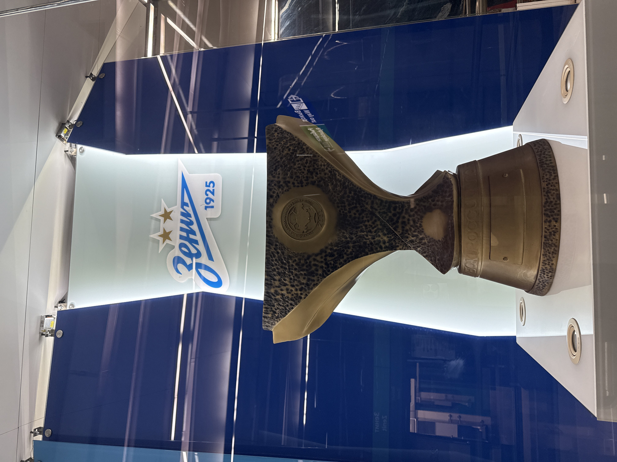 The image shows a trophy on display in what appears to be a museum or exhibition. The trophy is metallic, likely bronze or brass, with a textured surface and an ornate design. A circular emblem is visible on the front of the trophy. The backdrop consists of a white panel with the logo of Zenit St. Petersburg, a football club. The logo features the name "Zenit" in Cyrillic script, along with the year 1925 and stars. Behind the white panel are blue vertical stripes, possibly glass or plexiglass. The location is likely Saint Petersburg, Russia, given the presence of the Zenit logo, which is a prominent symbol of the city. The trophy itself is likely a prize or award related to Zenit football club. The display case appears modern with integrated lighting.
