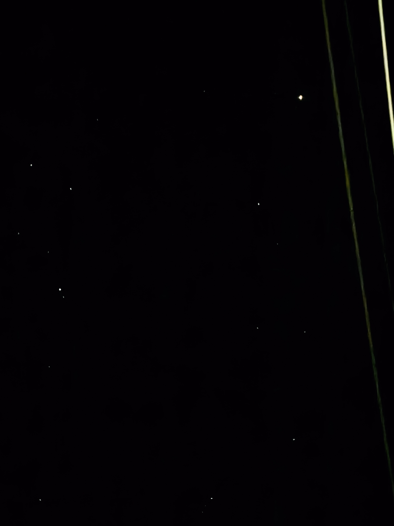 The image depicts a dark night scene in an unknown city. Several small, faint points of light, possibly stars, are scattered across the black background. On the right side of the image, there are a few indistinct, vertical lines, possibly wires or poles. The location is unspecified.