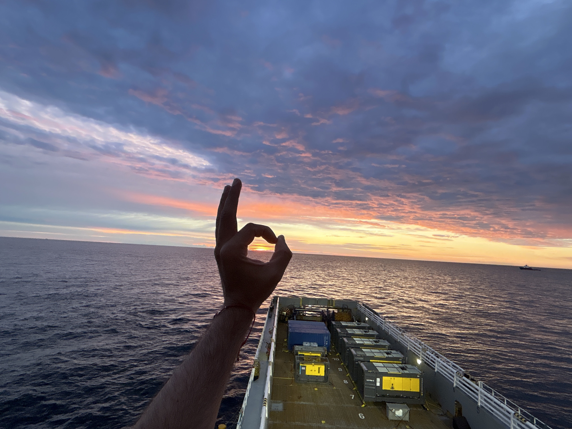 A person's arm and hand are in the foreground, making an "okay" hand gesture. The fingers are positioned to frame a portion of the setting sun over the ocean. The hand is on the left side of the image, extending from the bottom left corner upwards towards the center. The rest of the image shows a cargo ship at sea during sunset. The sky is filled with dramatic clouds colored in shades of pink, orange, and purple. The ocean is dark blue with visible waves. The deck of the cargo ship is visible in the lower right portion of the image, laden with large containers. In the distance, another ship can be seen on the horizon. The image appears to be taken from the deck of the cargo ship.