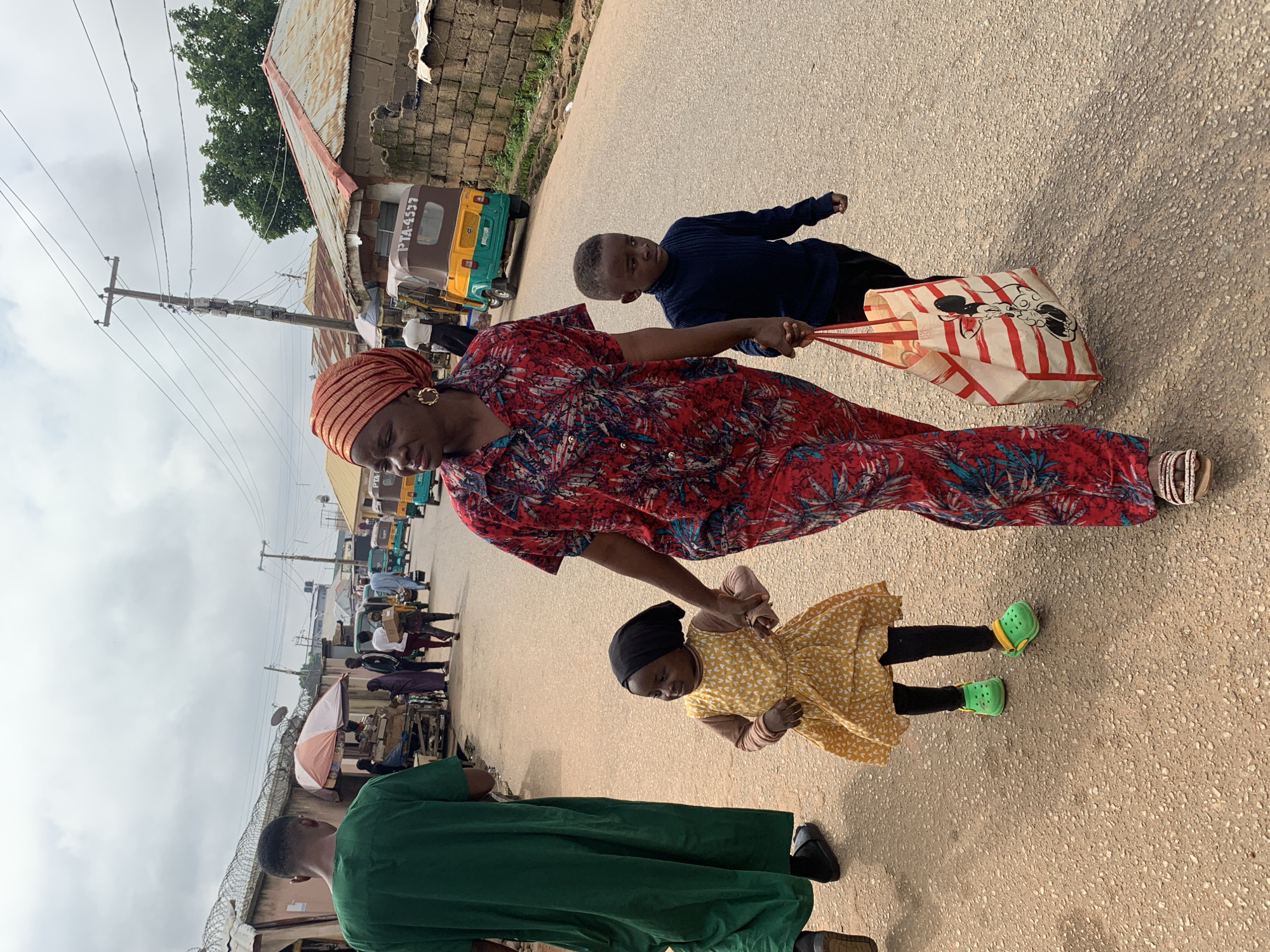 In Jos, Nigeria, a woman wearing a red and blue patterned dress and an orange headscarf walks down a dusty, paved road. She holds hands with a young boy in a dark blue sweater and black pants, who carries a red and white striped bag with a Mickey Mouse design. To her left, she also holds the hand of a young girl in a yellow dress with a white pattern and black leggings, wearing green clogs. A man in a green robe walks ahead of them on the left side of the road. In the background, several shops and stalls line the road, with a few auto-rickshaws, known as "Keke Napep," and other vehicles present. The sky is overcast with clouds.