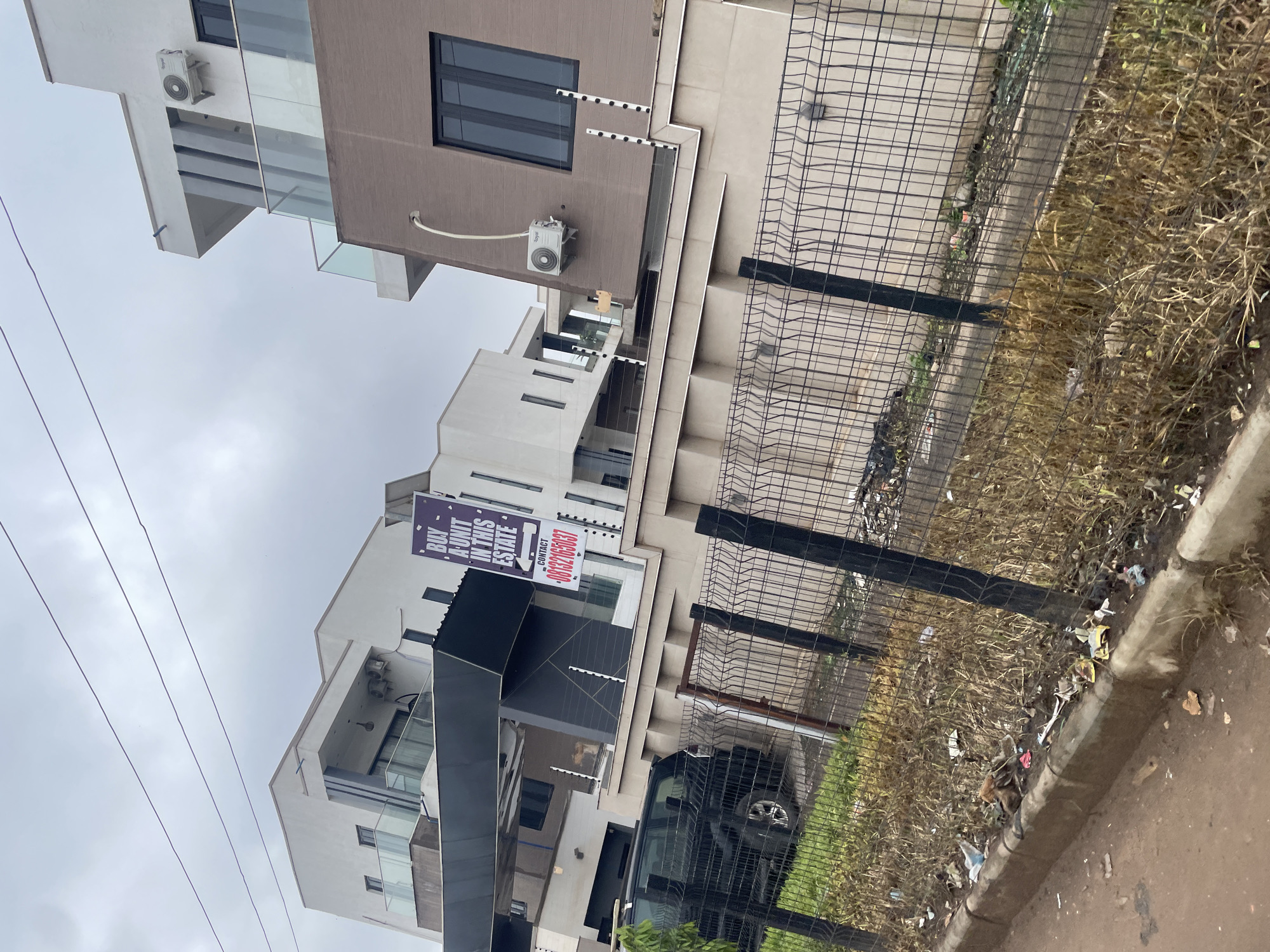This is a photograph of modern, multi-story residential buildings in Guzape, Nigeria, under an overcast sky. The buildings feature sleek designs with white and brown facades, glass balconies, and prominent air conditioning units. A dark-colored SUV is parked in front of one of the properties. A black metal fence with a barbed wire topping surrounds the property. A sign on the fence advertises units for sale with a contact phone number. The foreground shows a roadside with dry grass, dirt, and scattered litter. The overall impression is one of new construction in a developing urban area, with a clear contrast between the upscale residences and the less maintained roadside environment.