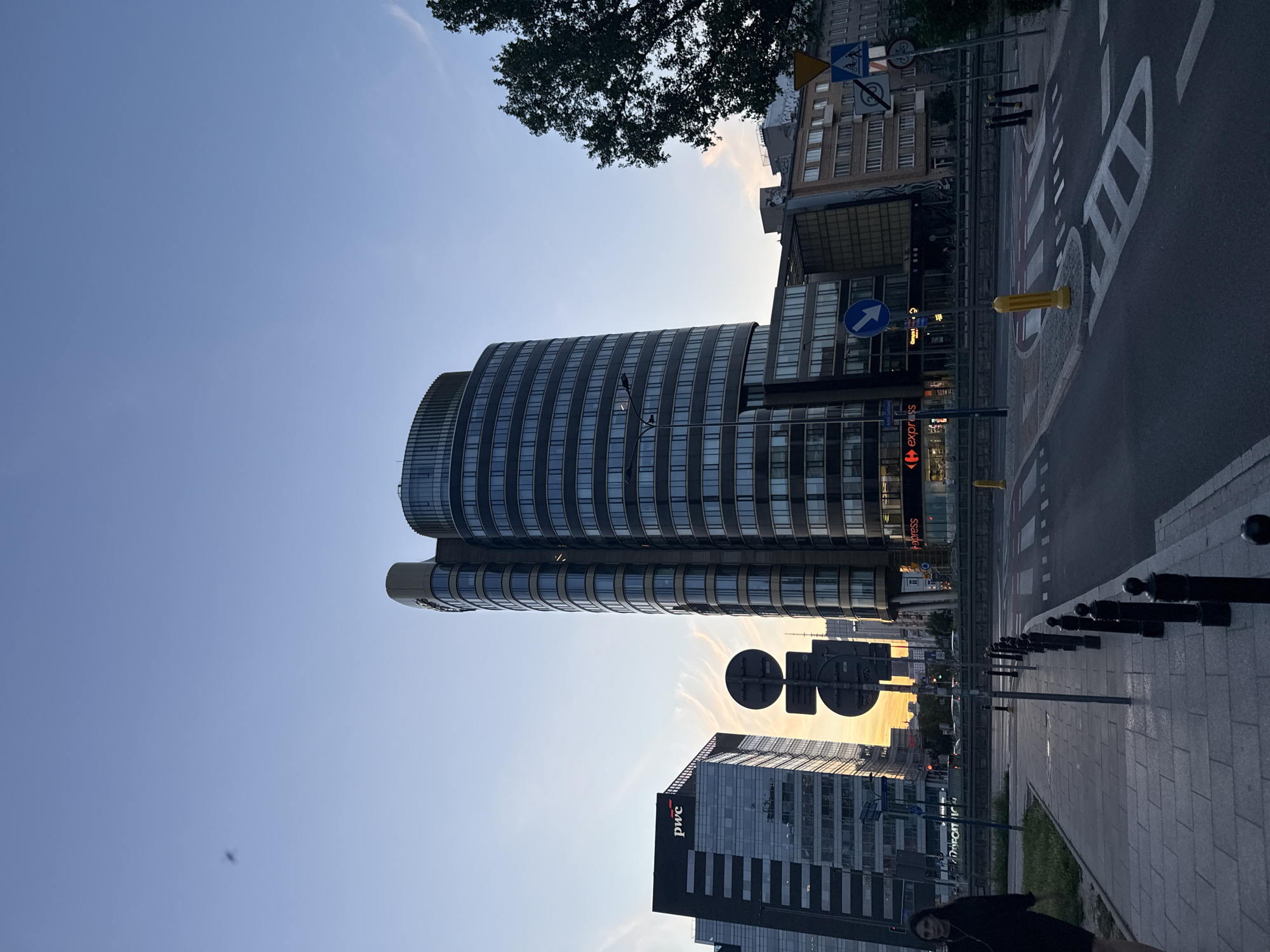 A vertical, low-angle shot captures a modern city street scene at dusk. Dominating the frame is a striking, curved glass building with horizontal bands of windows, reflecting the soft, warm light of the setting sun. To its right, a darker, rectangular glass-faced building displays the "pwc" logo in white.
The street in the foreground is paved with grey tiles, bordered by black bollards. A road with painted white lines and a bicycle lane marking is visible. Several traffic signs and streetlights dot the scene. A lone tree with dark green foliage is silhouetted against the pale blue sky.
The sky is mostly clear, with wispy clouds catching the golden hues of sunset. The overall mood is one of urban tranquility and the transition from day to night. The time of day is clearly dusk, with the sun low on the horizon and casting long shadows. The presence of modern architecture, street signs, and a bicycle lane suggests an urban environment, likely a business district. No specific emotions are overtly conveyed, but the soft lighting and calm atmosphere evoke a sense of peace.
Visible text includes "pwc" and "express" on building signage.