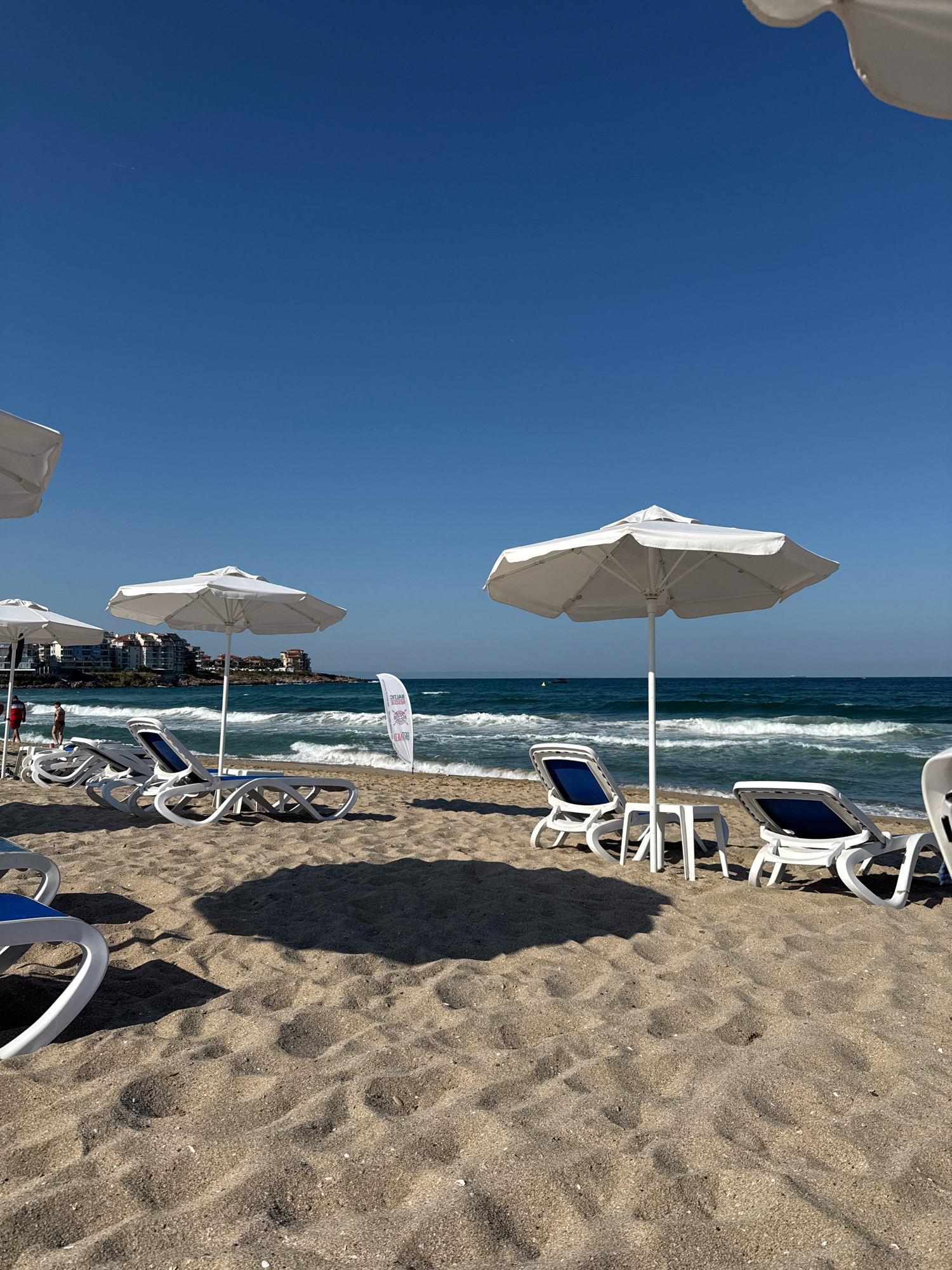 This is a photograph of a beach in Sozopol, Bulgaria, on a clear, sunny day. The foreground is dominated by sand, with shadows cast by the sun and beach umbrellas. In the middle ground, several white lounge chairs with blue cushions are arranged on the sand, some with small white tables. Three white beach umbrellas are visible, providing shade. A white flag with a red emblem is planted in the sand near the water's edge.
The background features the sea with moderate waves and a clear blue sky. Along the coastline, buildings with red-tiled roofs are visible, suggesting a developed area. Two figures are faintly visible walking on the beach in the distance. The overall scene depicts a typical seaside resort environment ready for visitors.
