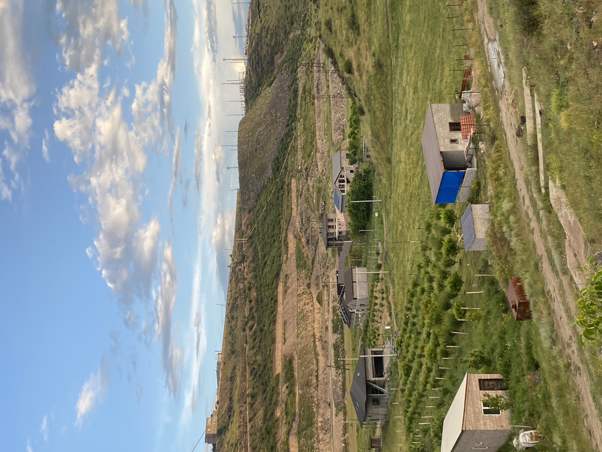 The image depicts a rural or semi-rural landscape near the city of Ptghni, Armenia, under a partly cloudy sky. In the foreground, a small, light-colored brick building with a white roof and a dark door is visible on the left. Adjacent to it is a rusty metal tank. A dirt path winds through the grassy terrain. Further into the mid-ground, there are several unfinished or under-construction residential buildings. One prominent structure has a gray roof and is partially covered by a blue tarp on one side, with exposed brickwork and construction materials visible. Another building to its left is also under construction, with visible structural elements and roofing. A small, rectangular structure with a light-colored roof is situated behind the building with the blue tarp. Beyond these buildings, there is a field with rows of young trees, possibly an orchard, separated by thin vertical posts.
In the background, a hillside rises, characterized by terraced levels of earth and sparse vegetation. Several power lines and utility poles are visible ascending the hill, and a cluster of taller towers, possibly communication or energy infrastructure, stands at the crest of the hill. The overall scene suggests a developing area with ongoing construction and a connection to the natural environment. There are no people or animals immediately visible in the image.