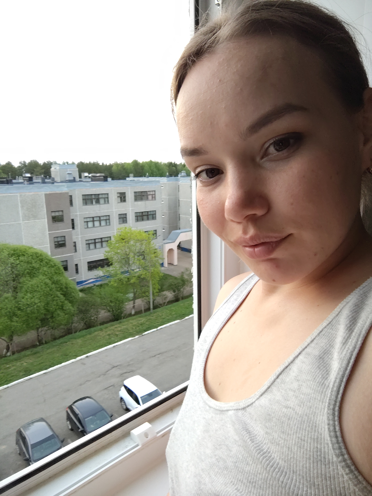 A young woman stands indoors near a window, taking a selfie. She wears a grey tank top and has her hair pulled back. Outside the window is a view of a multi-story building in the distance, along with greenery and three cars parked along a road. The cars are a dark gray, a black, and a white model. Based on the objects in the background, this is located in the city of Марковский, Russia.