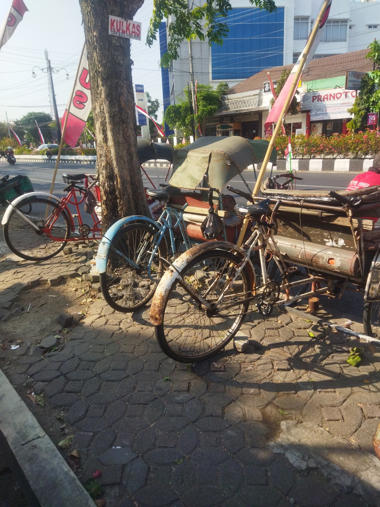 The image captures a sunlit street scene in Sukoharjo, Indonesia, featuring several parked bicycles and a pedicab. In the foreground, a weathered pedicab with a green canopy is the central object, its wooden bench and metal frame showing signs of age. Two bicycles are parked beside it: a red bicycle to the left and a blue one to its right. A third bicycle, white and rusty, is partially visible on the far right. The bicycles and pedicab are positioned on a paved sidewalk with a distinctive interlocking stone pattern. Behind the sidewalk, a street is visible with traffic, including a motorcycle. Trees and various buildings line the street, with a prominent building displaying a blue glass facade in the background. Red and white flags are planted along the roadside and on poles, suggesting a local event or celebration. A sign reading "SERBU KULKAS" is attached to a tree trunk. The overall impression is of a quiet, warm day in a bustling Indonesian city.