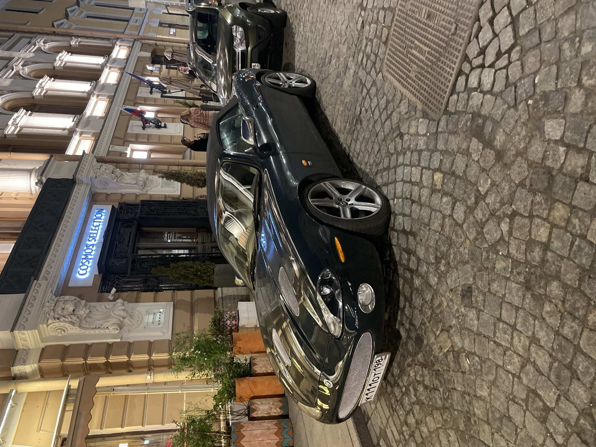 The image shows a dark green Aston Martin parked on a cobblestone street in Saint Petersburg, Russia. The car has a visible license plate: K111OT 198. In the background is a building with a sign that reads "COSMOS SELECTION" in neon blue. There are people standing near the entrance of the building, and Russian flags are displayed on the facade. The setting appears to be evening.