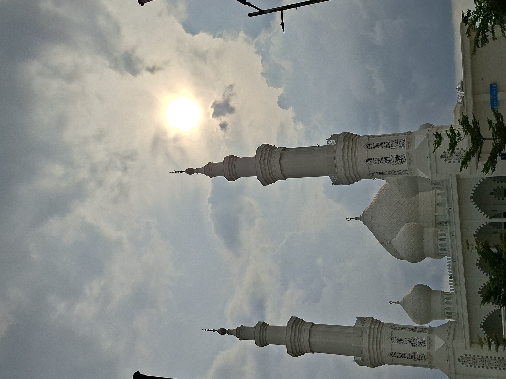 The image shows the facade of a white building with Islamic architecture, possibly a mosque, angled against a cloudy sky with the sun visible. The building features minarets, domes, and decorative latticework. The location is Ranto, Indonesia. There are trees partially visible on the right side of the frame. A light pole is visible in the upper central area of the picture.