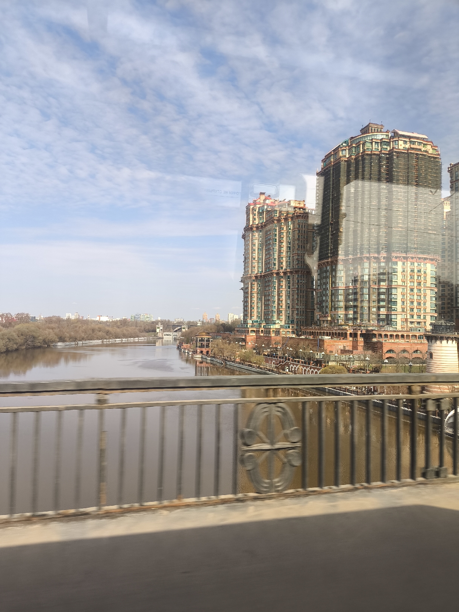 The image, captured through a window, presents a view of Khimki, Russia. A river runs through the scene, reflecting the pale sky with scattered clouds. The river's water is brown, and the opposite bank is lined with leafless trees. In the background are distant buildings, partially obscured by the haze and trees.
Dominating the right side of the frame are several tall, modern buildings. The construction netting obscures one of these buildings, implying an ongoing or recent construction project. The structures feature multiple tiers, each lined with windows, and topped with reddish roofing. The architectural design incorporates arched elements near the riverbank.
In the foreground, a metal bridge railing is visible. The railing is composed of vertical bars and decorative circular designs. The foreground area suggests the scene is being viewed from inside a vehicle.