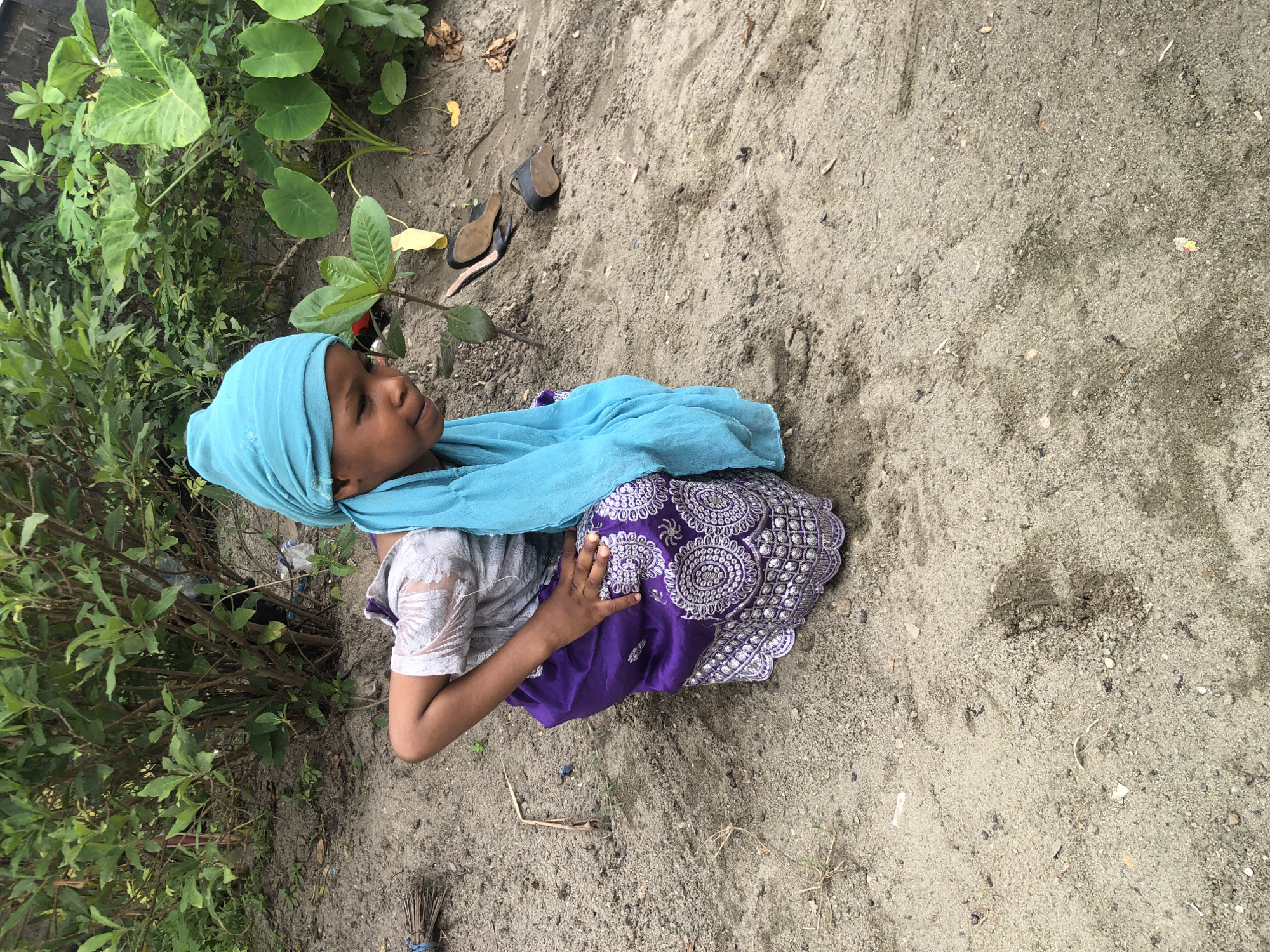 A young girl is crouching on sandy ground in Ado, Nigeria. She is wearing a light blue headscarf and a light grey shirt with white detailing. Her skirt is purple with a white embroidered pattern. She is looking down towards the ground with a slight smile. In the background, there are green leafy plants and some debris. A pair of black heeled shoes are lying on the ground to the right of the girl.