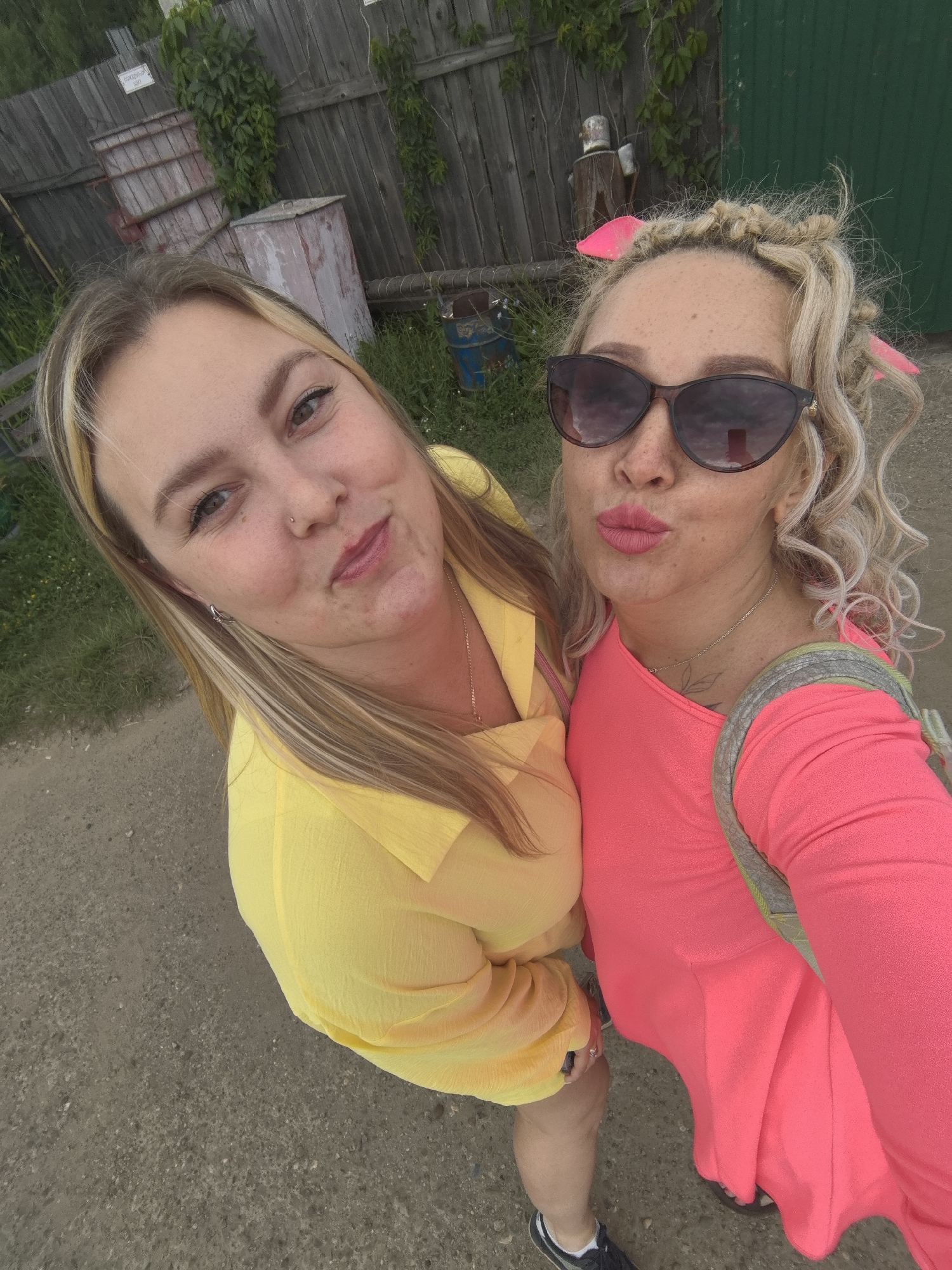 Two women are posing for a selfie outdoors in Noviy, Russia. The woman on the left has blonde hair with highlights and is wearing a bright yellow shirt. She is looking at the camera and smiling. The woman on the right has curly blonde hair styled with pink accessories and is wearing large, dark sunglasses and a bright pink top. She is puckering her lips as if for a kiss, also looking at the camera. A portion of a grey gravel path is visible in the foreground, with green grass and vegetation on either side. Behind the women, there is a weathered wooden fence, and various objects including what appear to be large wooden crates or bins, and a green structure in the far right. The lighting suggests it is daytime. There is some text visible on a sign attached to the fence, but it is too blurry to read.