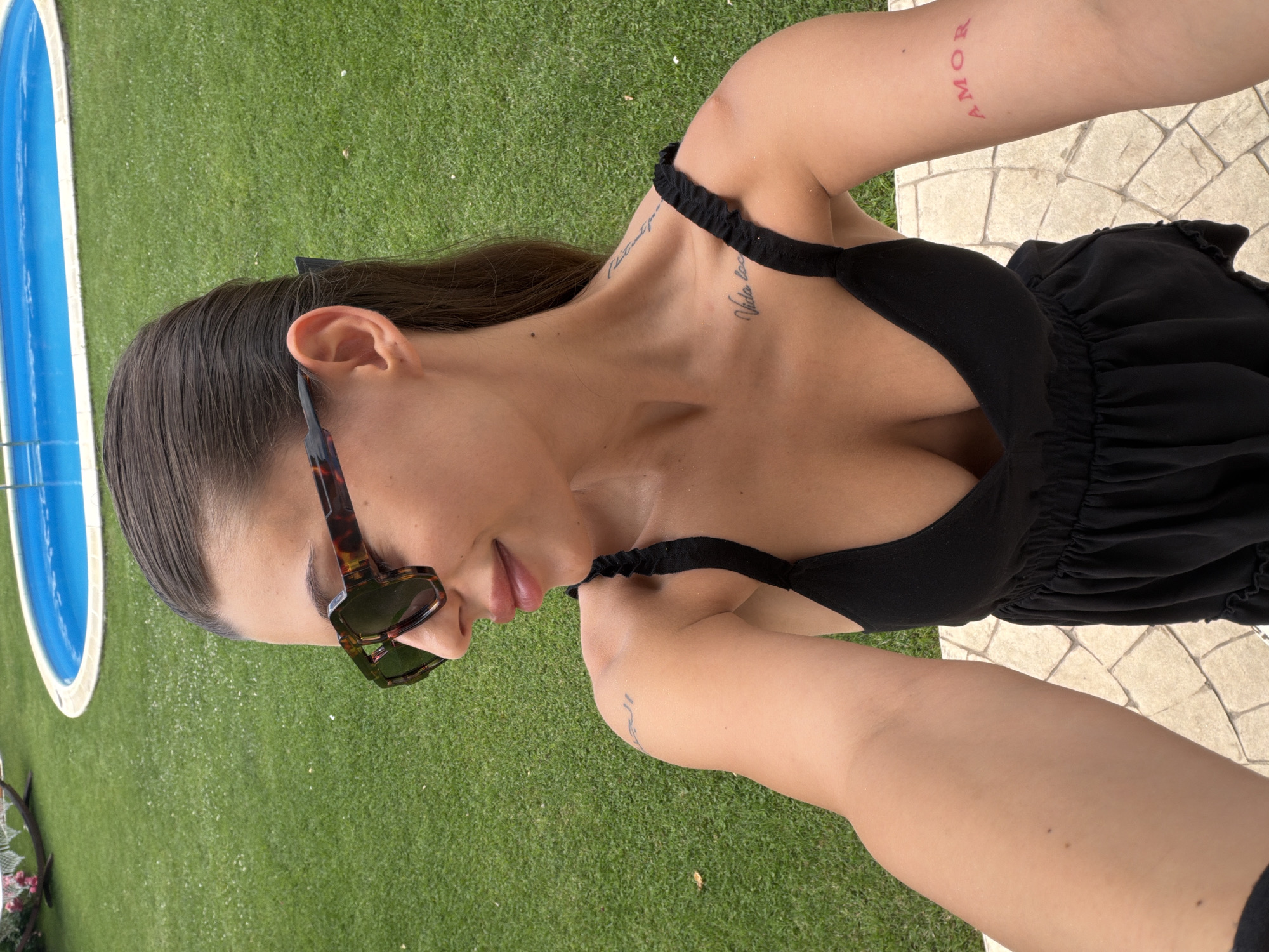 A young woman with dark hair pulled back, wearing sunglasses and a black dress, takes a selfie outdoors. She has tattoos on her left arm and shoulder, and a pink tattoo on her right forearm reads "AMOR". Behind her, a green lawn surrounds a blue swimming pool. The scene appears to be a private backyard, possibly in Aytos, Bulgaria, given the context provided, on a sunny day.