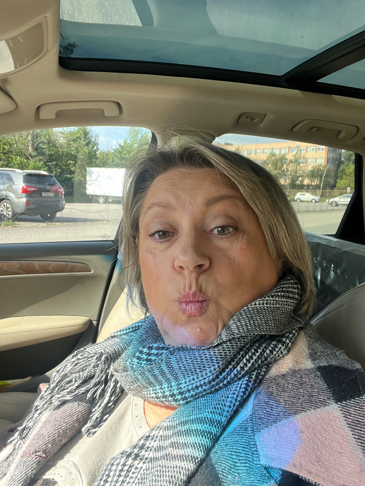 A middle-aged woman with blonde hair and blue eyes, wearing a gray and black patterned scarf and a tan shirt, is taking a selfie inside a car. She is puckering her lips as if for a kiss. The car has a sunroof, and through the windows, we can see trees, buildings, and other cars, suggesting an urban environment. The overall lighting is bright, indicating daylight. The interior of the car appears to be a light beige color. The woman is positioned in the driver's seat or the seat behind it, facing the camera directly.