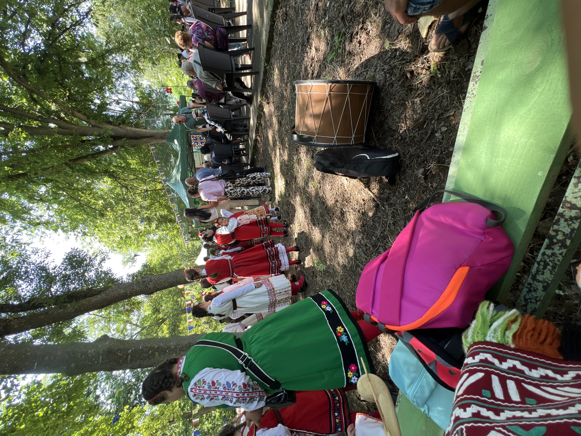 This image captures an outdoor cultural gathering in Bolyarovo, Bulgaria, under bright daylight. The scene is set in a park-like environment characterized by tall trees, dense green foliage, and sun-dappled ground with sparse dirt and fallen leaves.
In the immediate foreground, a green wooden bench holds a vibrant pink backpack with an orange zipper accent, a light blue fanny pack, and a maroon textile with a white pattern. To the right, a large, light brown drum with white lacing is resting on the ground beside a black instrument case. A person's lower leg and foot, wearing dark sandals, are visible on the far right, suggesting they are seated nearby.
The mid-ground is populated by numerous individuals. Several women and children are dressed in traditional Bulgarian folk costumes, featuring white embroidered blouses, colorful aprons, and long skirts in shades of green, red, and white. One woman, prominent on the left, wears a green apron dress with floral embroidery and a white embroidered blouse, carrying a black shoulder bag. Other costumed individuals are gathered behind her, some facing the main activity.
Further back, a diverse audience, including many older adults, is seated on black plastic chairs, facing a green canopy tent. A man in a light blue polo shirt and sunglasses stands near the tent, observing the crowd. Colorful bunting flags are strung between the trees, adding to the festive atmosphere. The background consists of more trees, with sunlight filtering through the canopy.