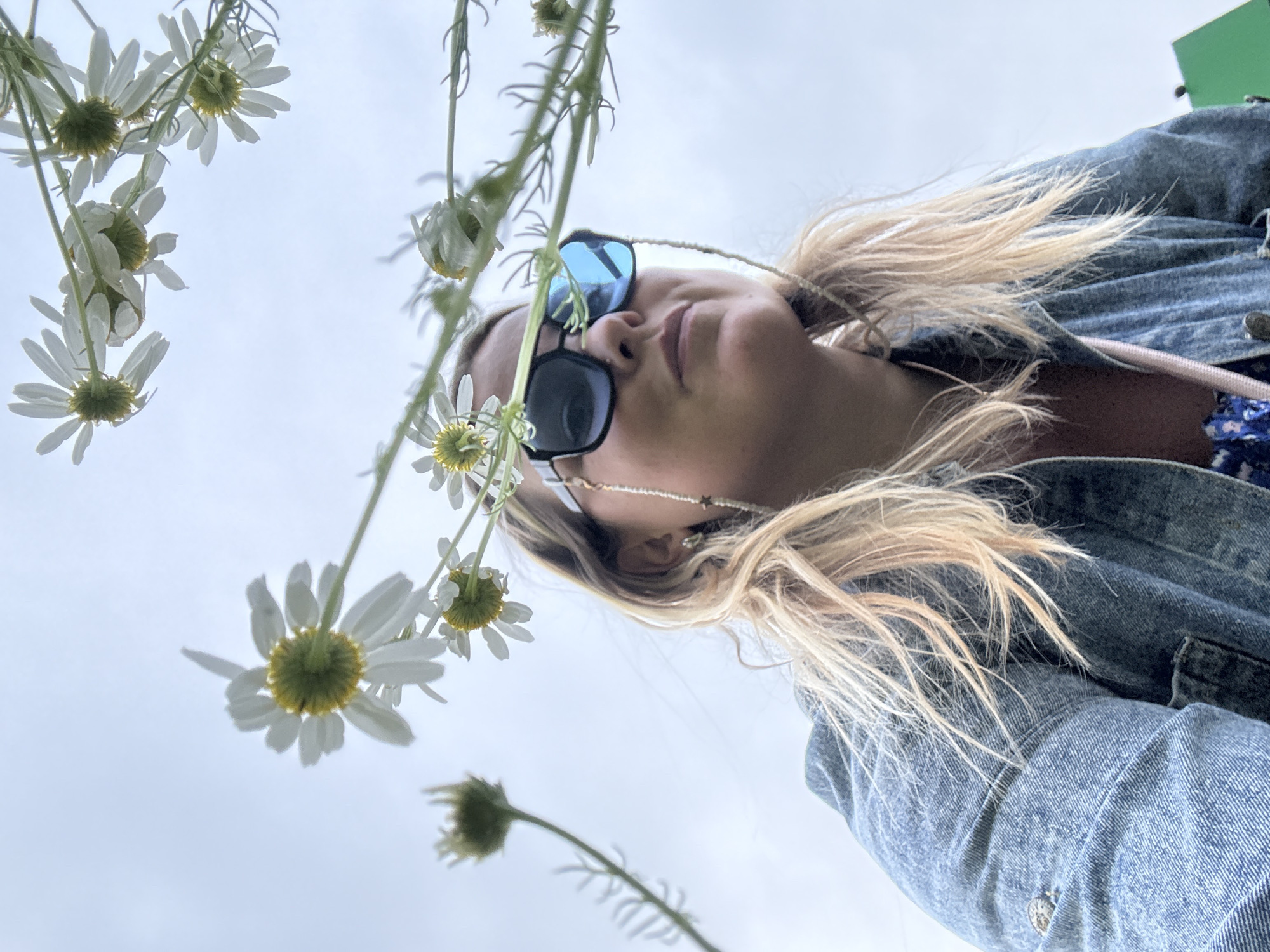 A low-angle, medium shot captures a fair-skinned woman with blonde hair, wearing large, dark-lensed sunglasses and a denim jacket, looking upwards with a neutral expression. She is positioned in the foreground, partially obscured by white daisies with yellow centers that are in focus, suggesting they are close to the camera. The background is a pale, overcast sky. The woman is wearing a beaded chain around her neck. A hint of a blue floral pattern is visible on her clothing beneath the jacket. The image does not contain any identifiable landmarks or features to suggest a specific location within Марковский, Russia.