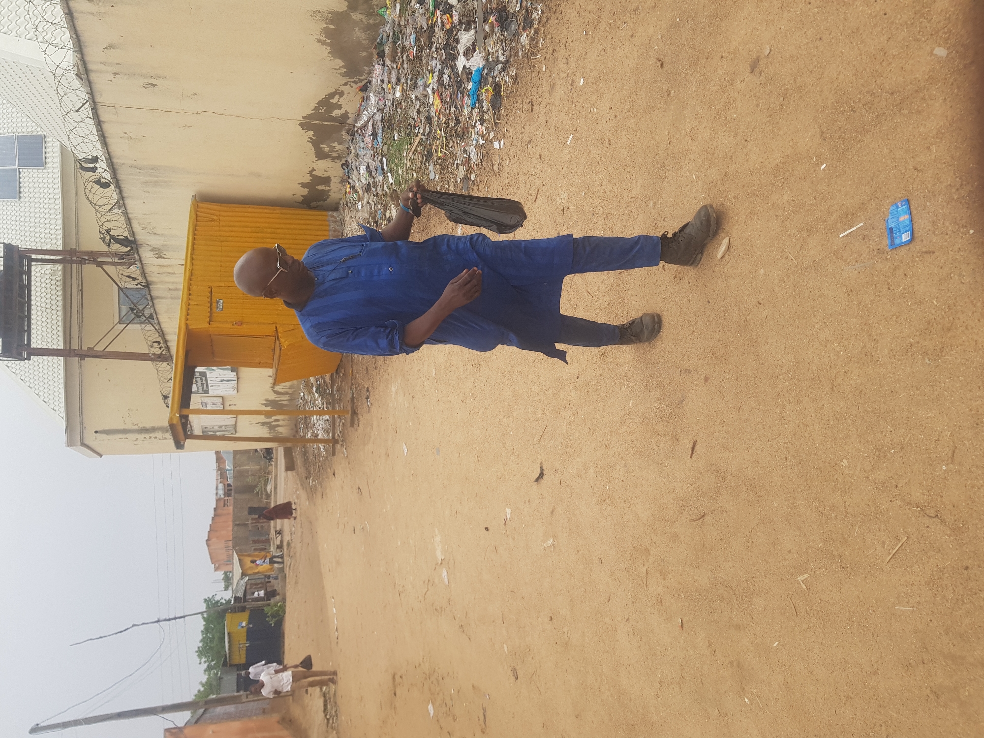 A medium shot captures a bald man in a blue traditional attire walking on a dirt road in Dabawa, Nigeria. He carries a black plastic bag. The road is strewn with debris. To his right, there is a large pile of trash against a wall. A yellow structure stands near the wall, with buildings visible in the distance. Overhead, there is an overcast sky.