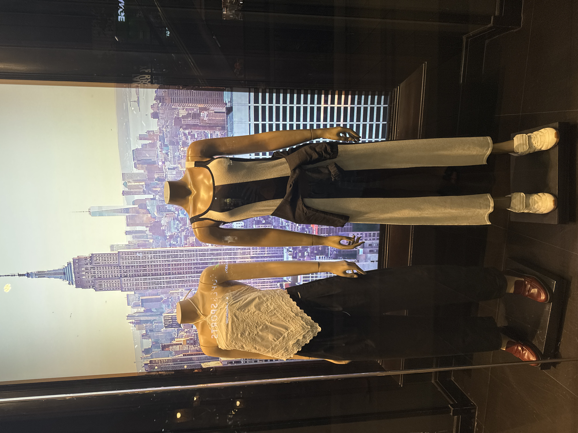 Two mannequins are displayed in a storefront window against a backdrop of the New York City skyline, including the Empire State Building and One World Trade Center. The top mannequin wears a gray and black dress with a black jacket tied around the waist and white sneakers. The lower mannequin is dressed in a white lace top and black pants, paired with brown loafers. The window frame is black. The location in บ้านบางรักษ์, Thailand is not applicable to this image.