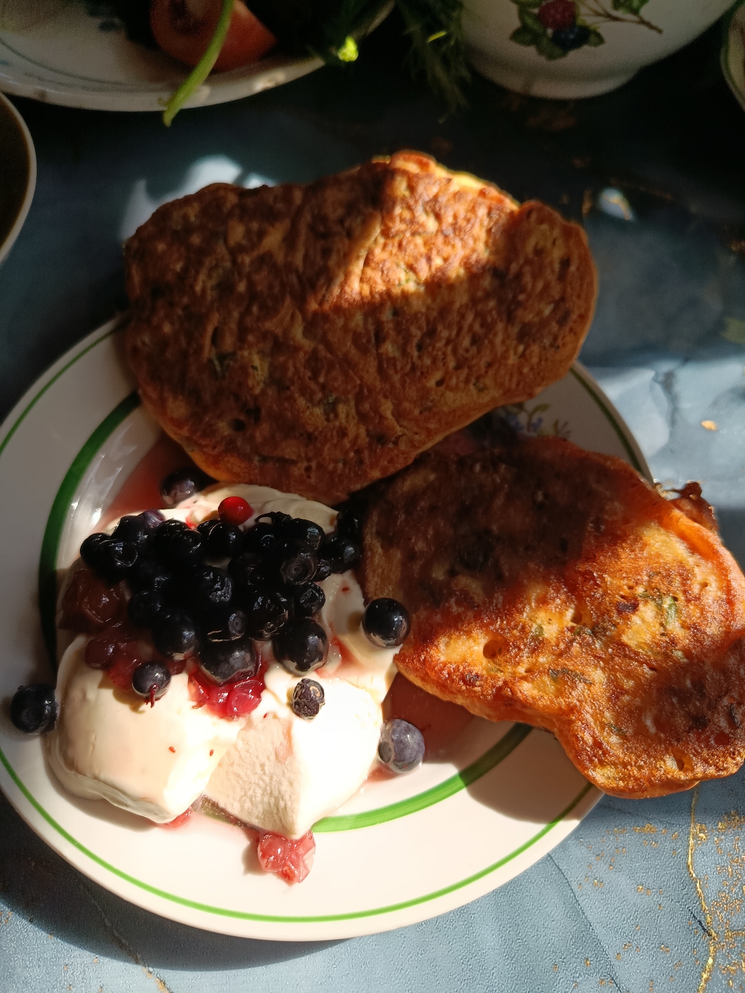In the city of Veraya, Russia, a close-up, high-angle shot captures a delectable breakfast spread. On a white plate with green trim, two golden-brown pancakes are arranged. One pancake rests atop the other, with a portion of the lower pancake visible. Nestled beside the pancakes, on the left side of the plate, is a generous scoop of creamy white dessert, possibly ice cream or yogurt, adorned with a vibrant assortment of fresh blueberries and a single red currant. A drizzle of reddish syrup, likely berry compote or sauce, flows around the dessert and onto the plate. The background is a softly blurred blue surface, possibly a table, with hints of sunlight casting dappled shadows and golden flecks, suggesting an outdoor or sunlit indoor setting.