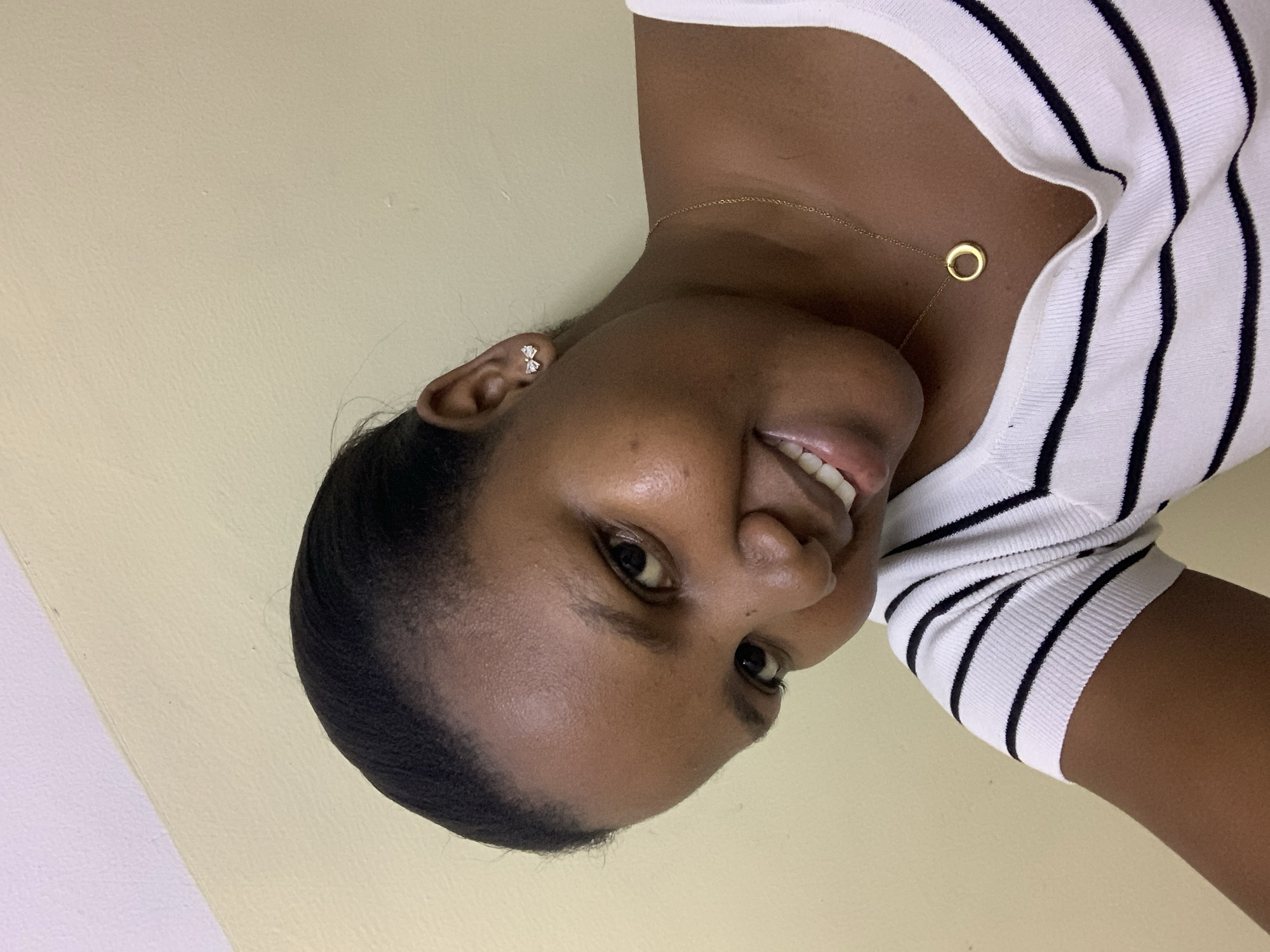 A young woman with dark skin and dark hair pulled back from her face is smiling. She is wearing a white shirt with black horizontal stripes and a thin gold necklace with a circular pendant. She has small stud earrings in her ear. The background is a plain, light beige or yellow wall. There is no information in the image to indicate that the location is Dar es-Salaam, Tanzania, or any other specific location.