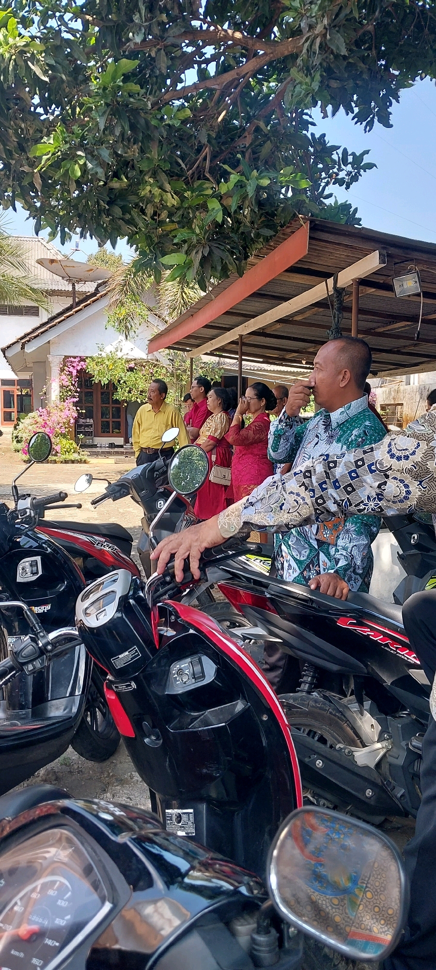 In Keling, Indonesia, a group of people stand near several parked motorcycles under a shaded structure. The man in the foreground, wearing a batik shirt, is drinking from a small cup. To his left, several women in traditional red and gold attire observe something to their left. A man in a yellow shirt stands further left, also looking in the same direction. The background reveals a white house with pink flowering bushes and a satellite dish on the roof. The environment suggests a warm, sunny day.