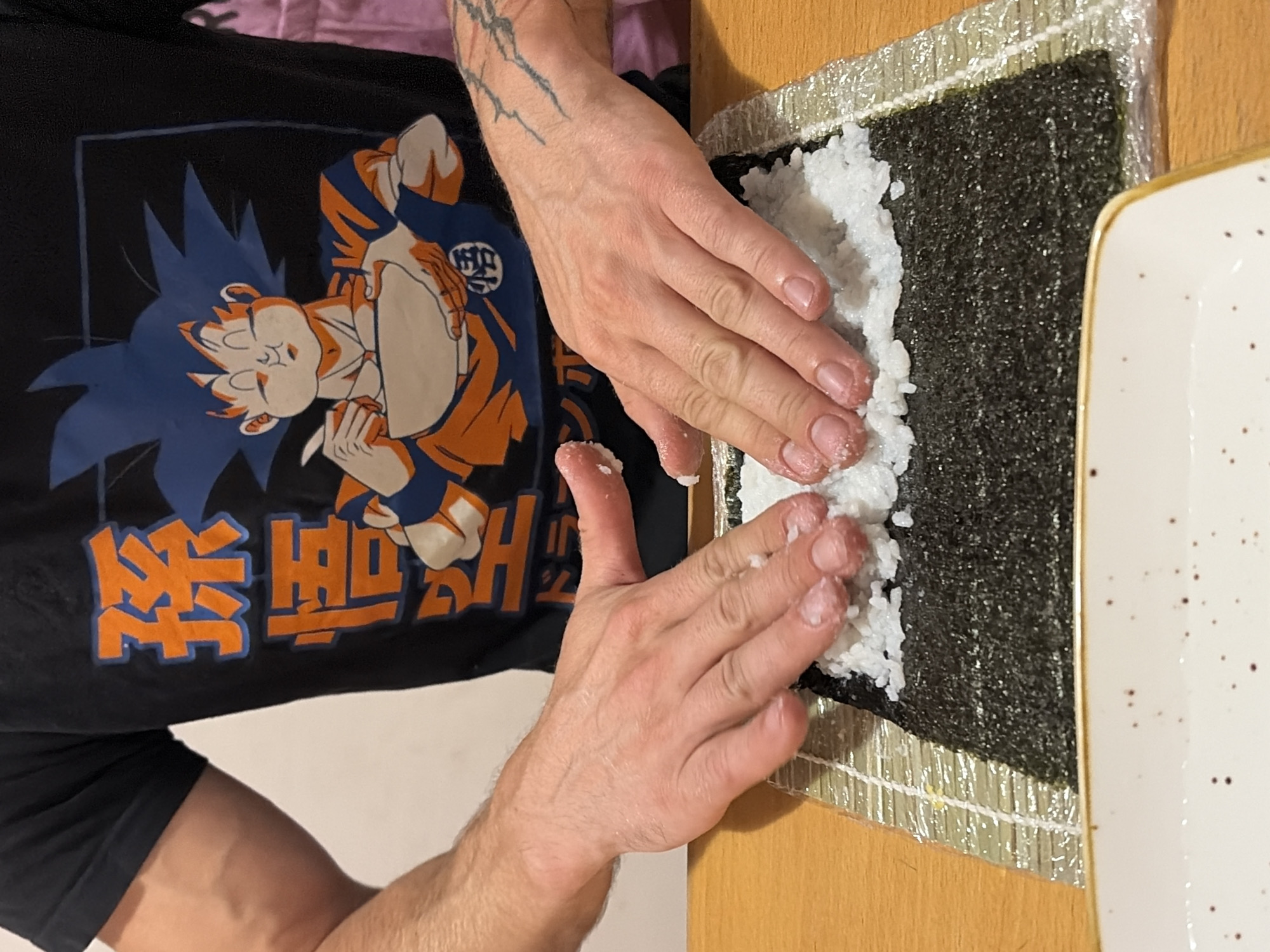 A person is making sushi. They are wearing a black t-shirt with an anime character on it. Their hands are pressing rice onto a sheet of nori seaweed, which is placed on a bamboo sushi mat. The sushi mat is on a wooden surface. In the foreground, there is a white plate with a gold rim. The background is a white wall. The person's left arm has visible veins and the right arm has a tattoo. The image does not provide any information about the location in the city of Budva, Montenegro.