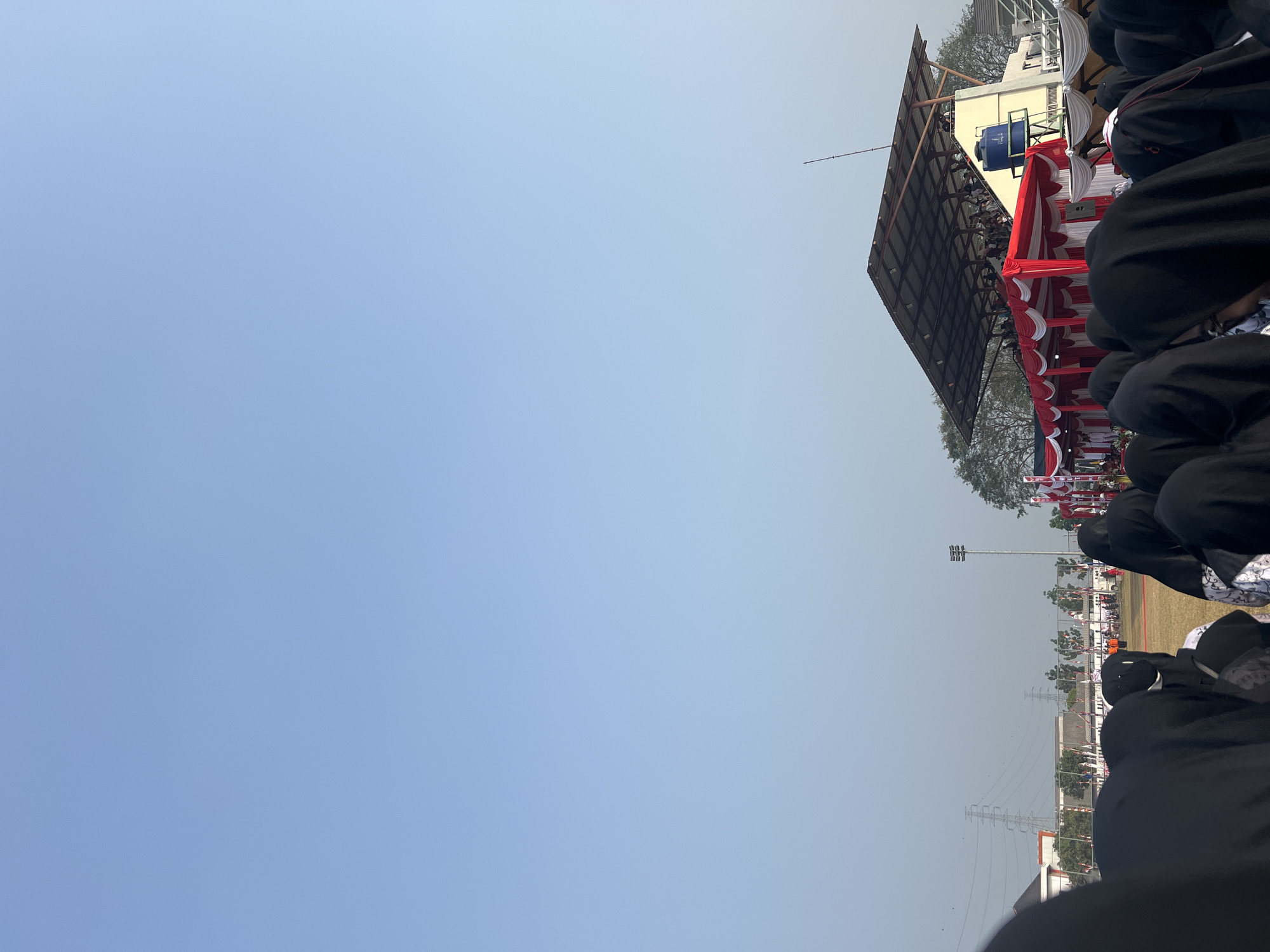 In Sukamantri, Indonesia, a large crowd of people wearing hijabs stands in the foreground, their backs to the camera. They are facing a stage decorated with red and white drapery, where many people are gathered, some seated under a covered roof. A blue water tank is visible on a yellow building behind the stage. In the background, transmission towers and buildings are seen under a clear blue sky. The ground is covered with dry grass, suggesting an outdoor event.