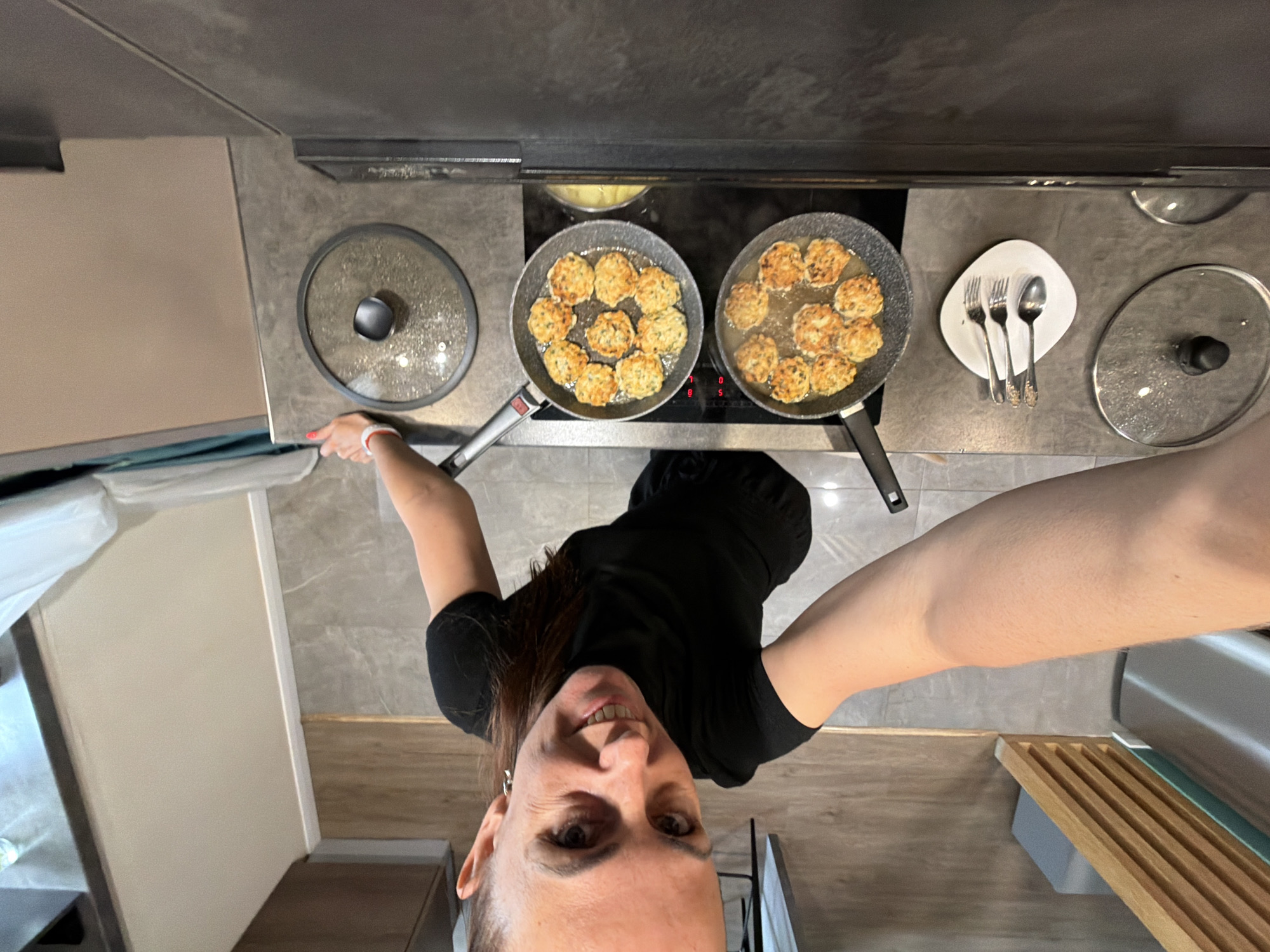 An overhead selfie captured in a modern kitchen shows a woman cooking. She is smiling directly at the camera, holding it high above her. She has dark hair, is wearing a black t-shirt, and has red nail polish visible on one hand.
The scene focuses on a stovetop with two frying pans actively cooking disc-shaped, golden-brown patties or fritters in oil. The oil in the pans is bubbling, indicating active cooking. On the dark gray countertop, next to the stovetop, are two pot lids – one clear glass with a black knob and another metal one. A white, irregularly shaped plate holds three pieces of silver cutlery (two forks and one spoon).
The kitchen features dark gray countertops and what appears to be a matching stovetop or induction hob, with faint digital numbers ("0", "5") visible on its surface. Above the cooking area, a dark range hood is visible. The flooring is a mix of wood laminate and gray tile, and the walls are light-colored. To the far right, a wooden slatted drying rack or mat is placed on the counter. The overall impression is a domestic setting where a meal is being prepared.