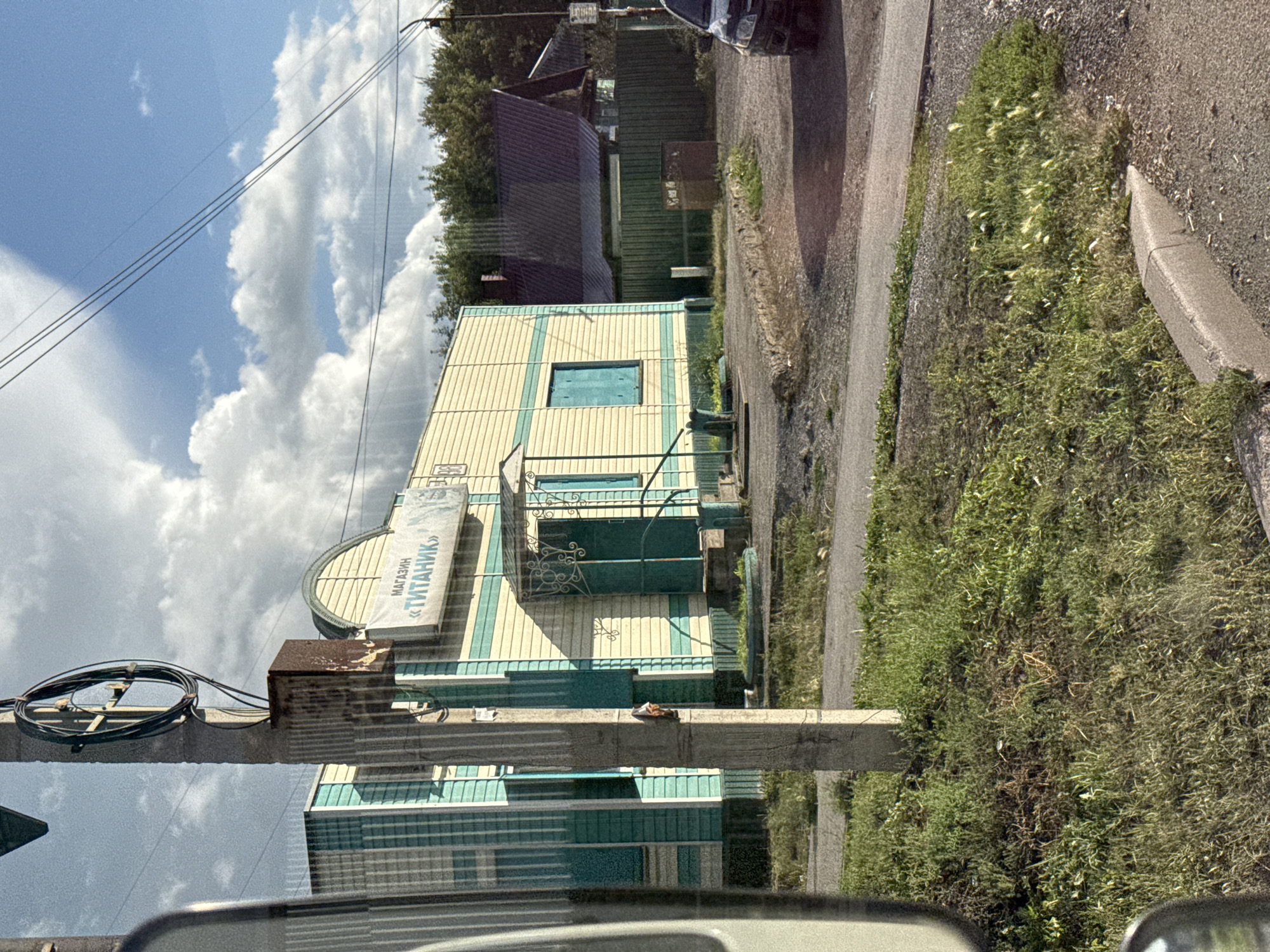 In an outdoor scene, a weathered concrete utility pole stands in the foreground, positioned in front of a light-colored building with teal trim. The building has a banner that reads "МАГАЗИН «ТИТАНИК»" in Russian, indicating it is a store named "Titanic". To the right of the building, a dark-colored sedan is parked on a gravel lot. Further to the right, a dark green fence encloses a property with trees visible beyond it. The sky above is a mix of blue and white clouds, with power lines crisscrossing the upper portion of the image. The ground in front of the building and pole is covered in patchy grass and gravel.