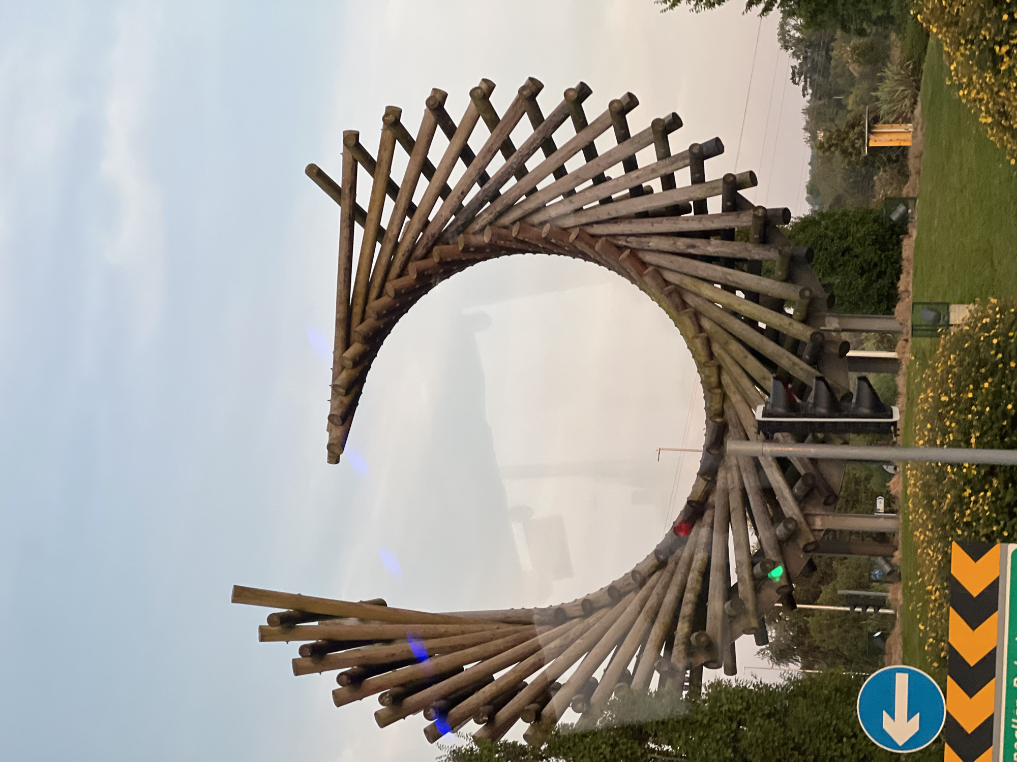The image shows a large, wooden, spiral sculpture at a roundabout in Letterkenny, Ireland. The sculpture is made of numerous wooden poles arranged in a curving, helical pattern, resembling a DNA double helix or a twisted ribbon. The sculpture is positioned in the center of the roundabout, with lush green grass and yellow flowers surrounding its base. A traffic light is visible near the sculpture, with the green light illuminated. A blue circular road sign with a white arrow pointing left is also present, indicating a turning direction. The background consists of trees, hills, and a pale sky. The overall impression is of a public art installation in a suburban or semi-rural setting.