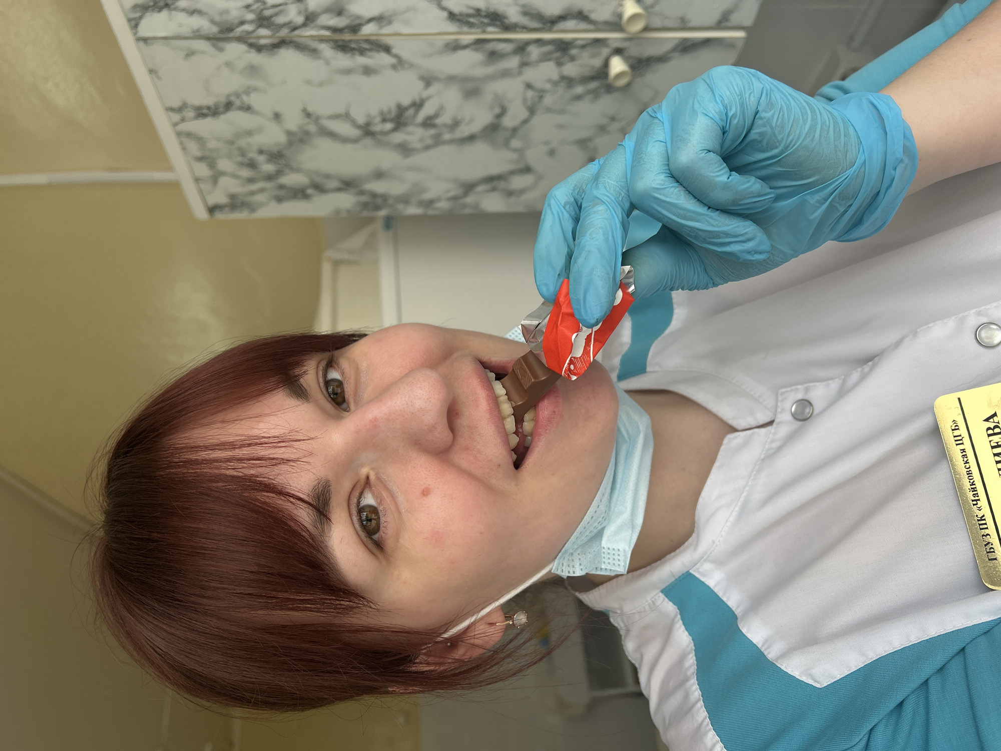 A mid-shot captures a person, likely a woman with reddish-brown hair and brown eyes, looking directly at the camera with a playful expression. She is wearing a white medical coat with light blue accents and light blue medical gloves. A light blue surgical mask is pulled down, resting under her chin. She is holding a partially unwrapped chocolate bar in a red and silver wrapper with her gloved hand, taking a bite from it.
A gold identification badge is clipped to her coat on the right side of the image. The setting appears to be an indoor professional environment, possibly a medical office or clinic, suggested by the uniform. The background consists of a plain light-colored wall on the left and a ceiling with a grey and white marbled pattern on the right, separated by a white trim. One ear is visible, adorned with a small, light-colored stud earring.
Visible text on the gold identification badge, written in Cyrillic, reads "ГБУЗ ПК «Чайковская ЦГБ»", which translates to "State Budgetary Healthcare Institution of Perm Krai 'Chaykovskaya Central City Hospital'". Below this, the surname "ЗУБИЕВА" (ZUBIEVA) is visible. The image conveys a moment of lightheartedness or a break during work.