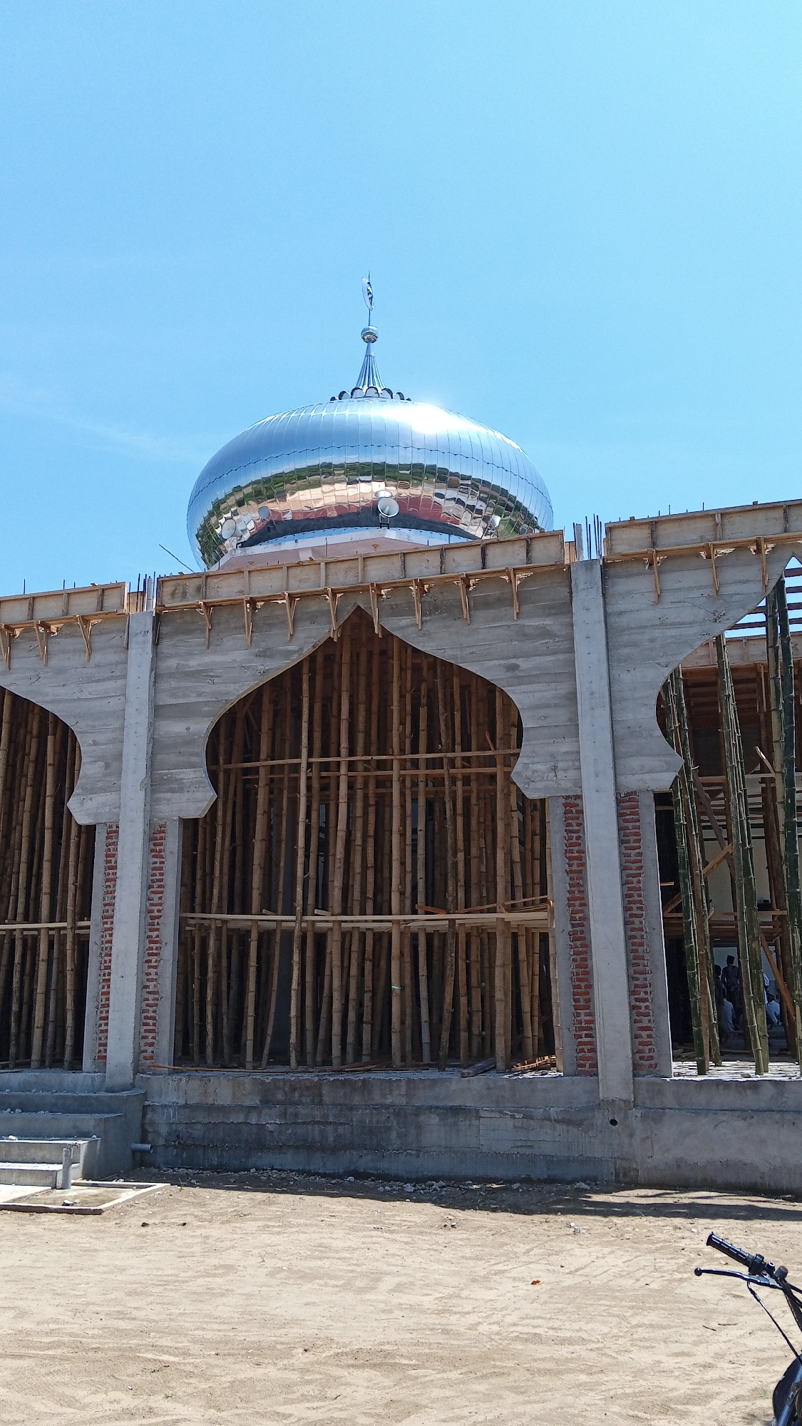 The image shows the exterior of a mosque under construction in Uteunreutoh, Indonesia. A large, polished silver dome is visible at the top, reflecting the clear blue sky. The building's facade is made of concrete with arched openings. Inside the openings, numerous vertical bamboo poles are arranged, likely as scaffolding or temporary support. Some brickwork is visible on the concrete pillars. In the background, through one of the openings, several people can be seen. The ground in front of the mosque is a sandy, dirt surface. A portion of a bicycle handlebar and mirror is visible in the lower right corner. The scene suggests ongoing construction work on a religious building.