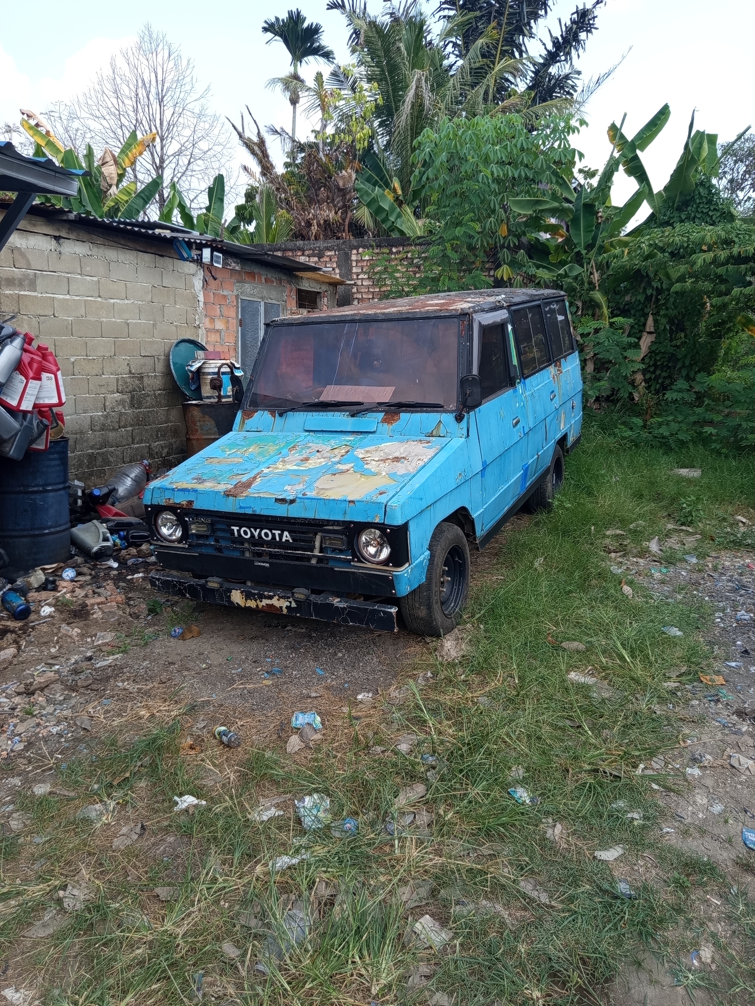 The image shows an abandoned, light blue Toyota van with peeling paint and rust. The van is parked in a cluttered outdoor area with overgrown grass and scattered debris. Behind the van is a brick building with a corrugated metal roof, and to the right are dense green trees and foliage, suggesting a tropical or subtropical environment. To the left of the van are several large plastic barrels and stacks of red plastic containers. The scene appears to be in a disused or informal settlement. The location is identified as Palembang, Indonesia.