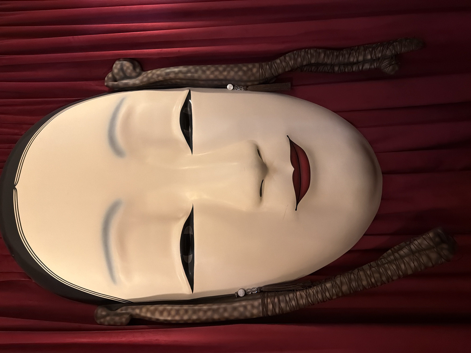 A close-up shot presents a Japanese Noh mask resting against a background of red draped fabric. The mask is oval with a pale complexion, narrow, closed eyes, a small nose, and painted red lips. The mask is flanked by what appears to be wrapped sticks, likely part of a presentation or display arrangement. No information about the location in Moscow, Russia, can be determined from the image.